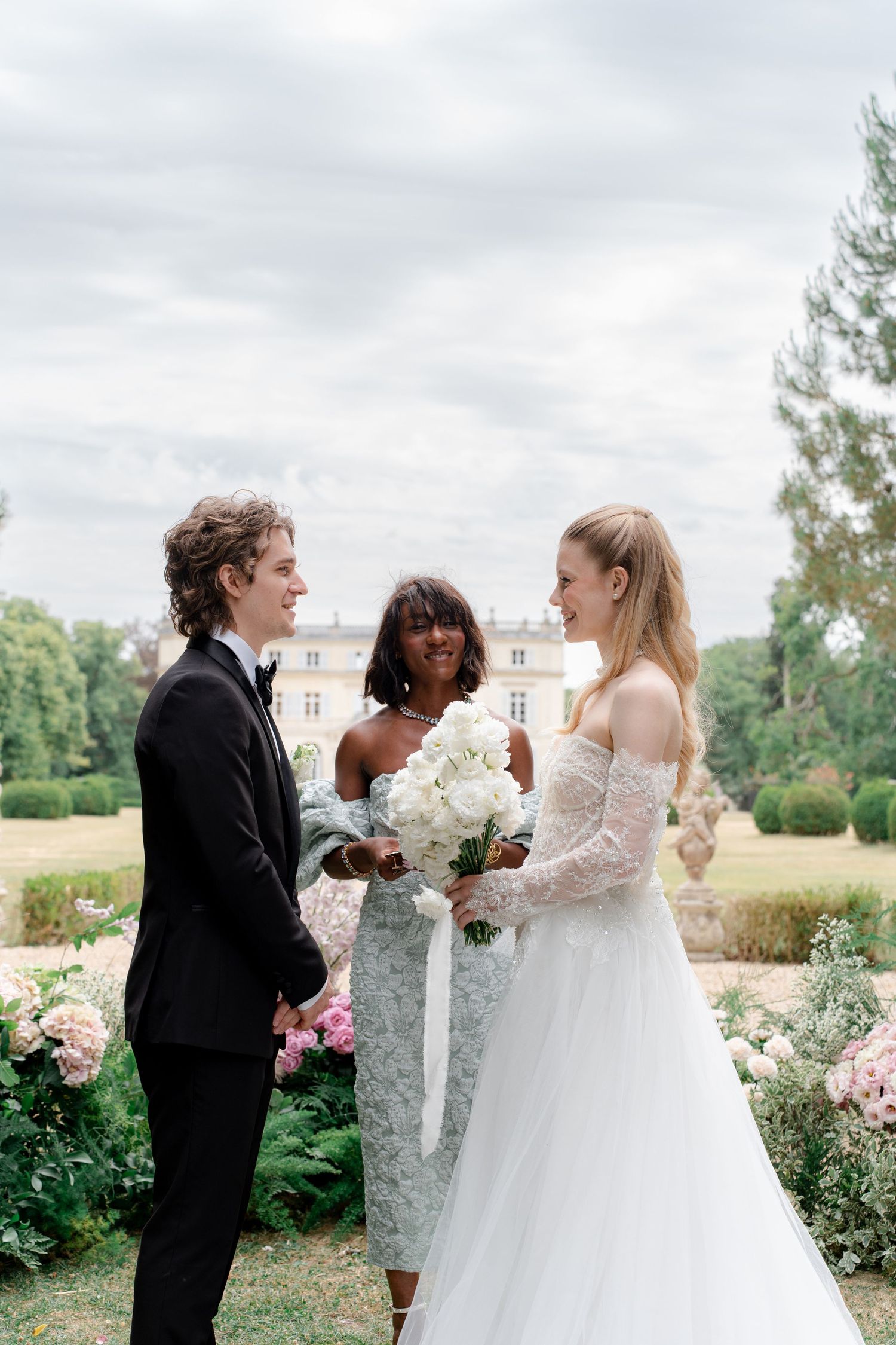 Elegant outdoor wedding ceremony with bride holding white bouquet, groom in tuxedo, and officiant at a French chateau.