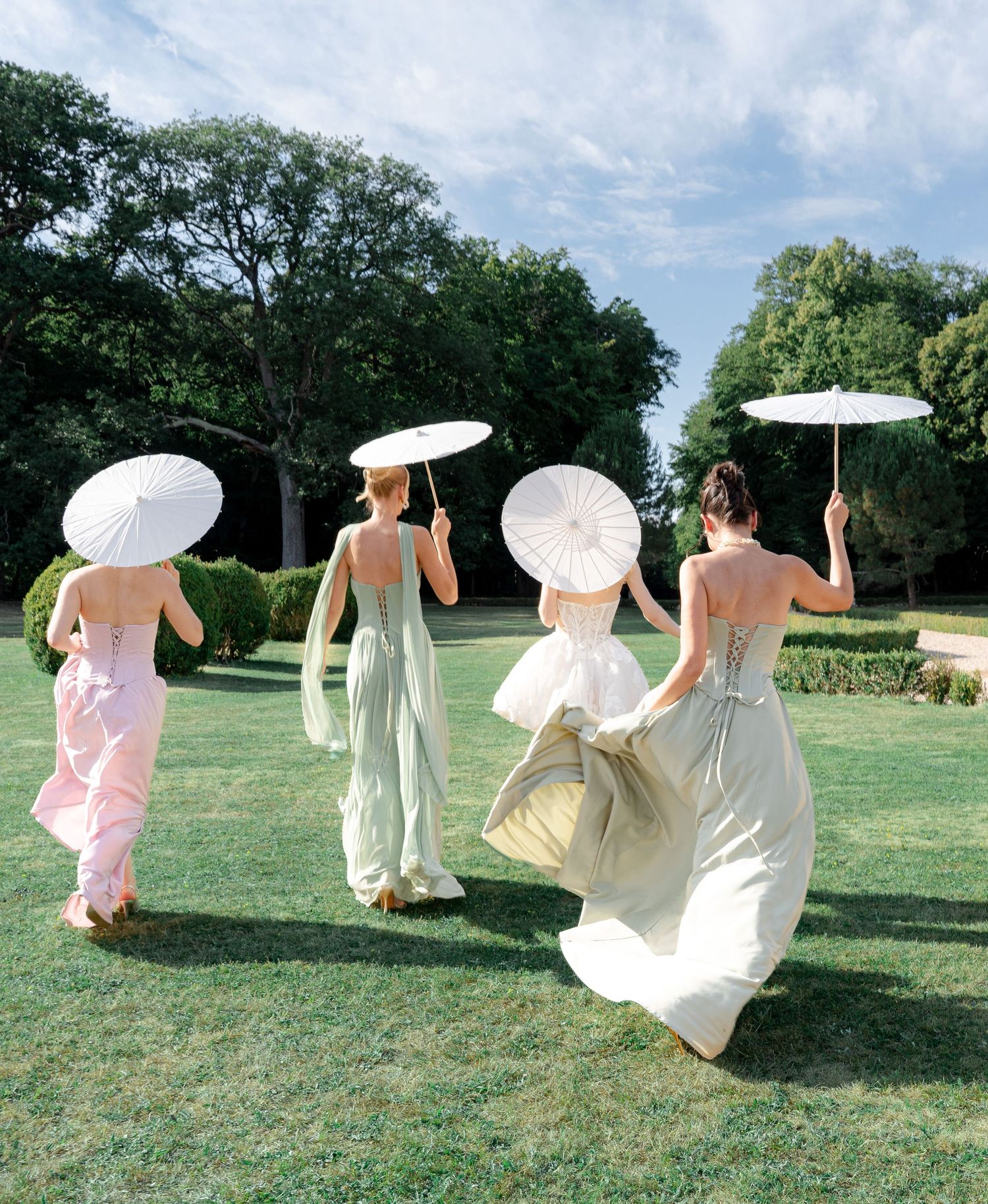 Four women in pastel gowns walk across green lawn holding white parasols, viewed from behind on a sunny day.