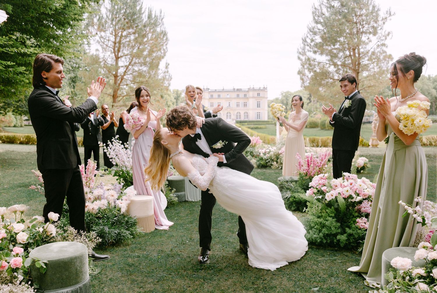 Bride and groom share romantic dip kiss surrounded by wedding party in lush garden with pink flowers and elegant chateau.