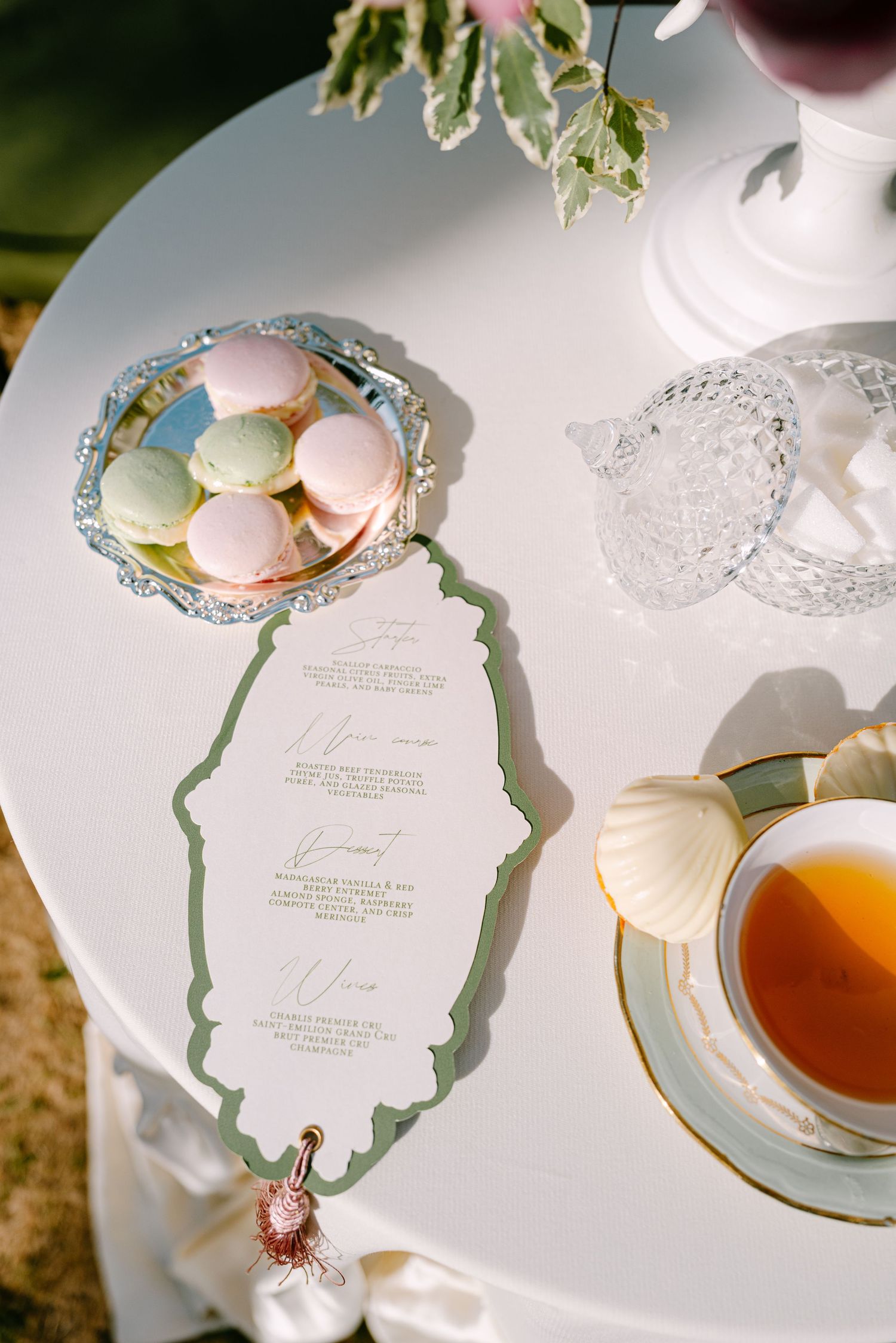 Elegant wedding menu card with green border beside macarons, tea cup, and crystal dish on a white table.