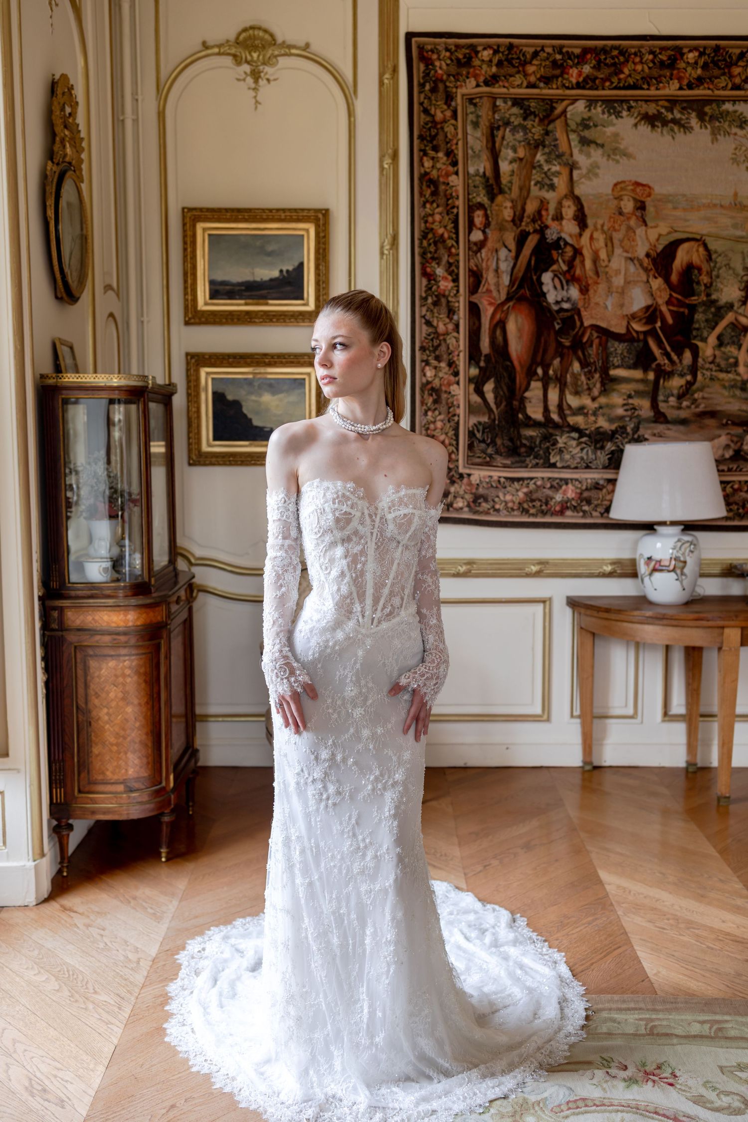 Bride in elegant lace wedding gown with long sleeves poses in ornate room with tapestry and gold-framed artwork.