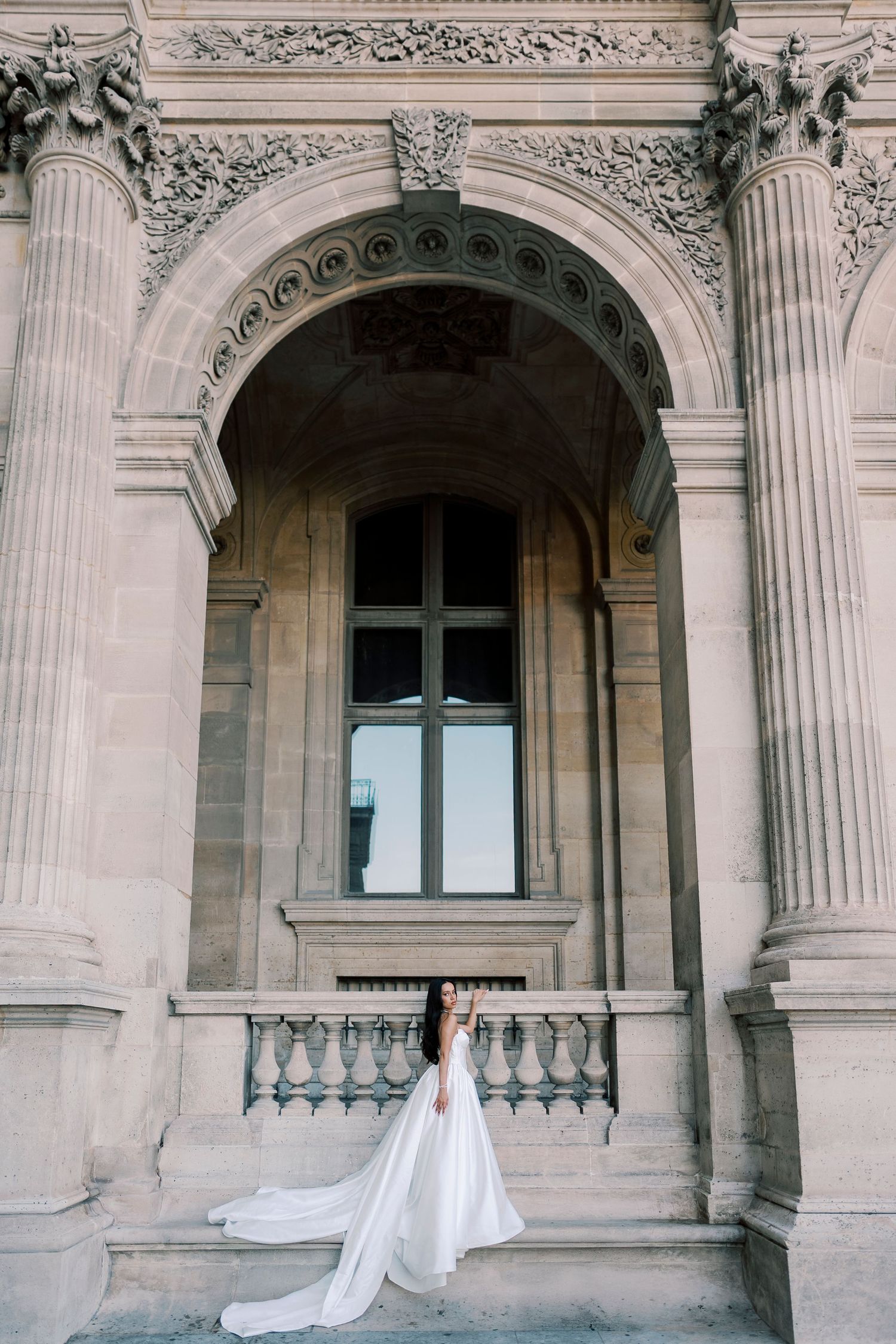 Bride in white gown with long train poses on steps of ornate classical stone building with arched window.