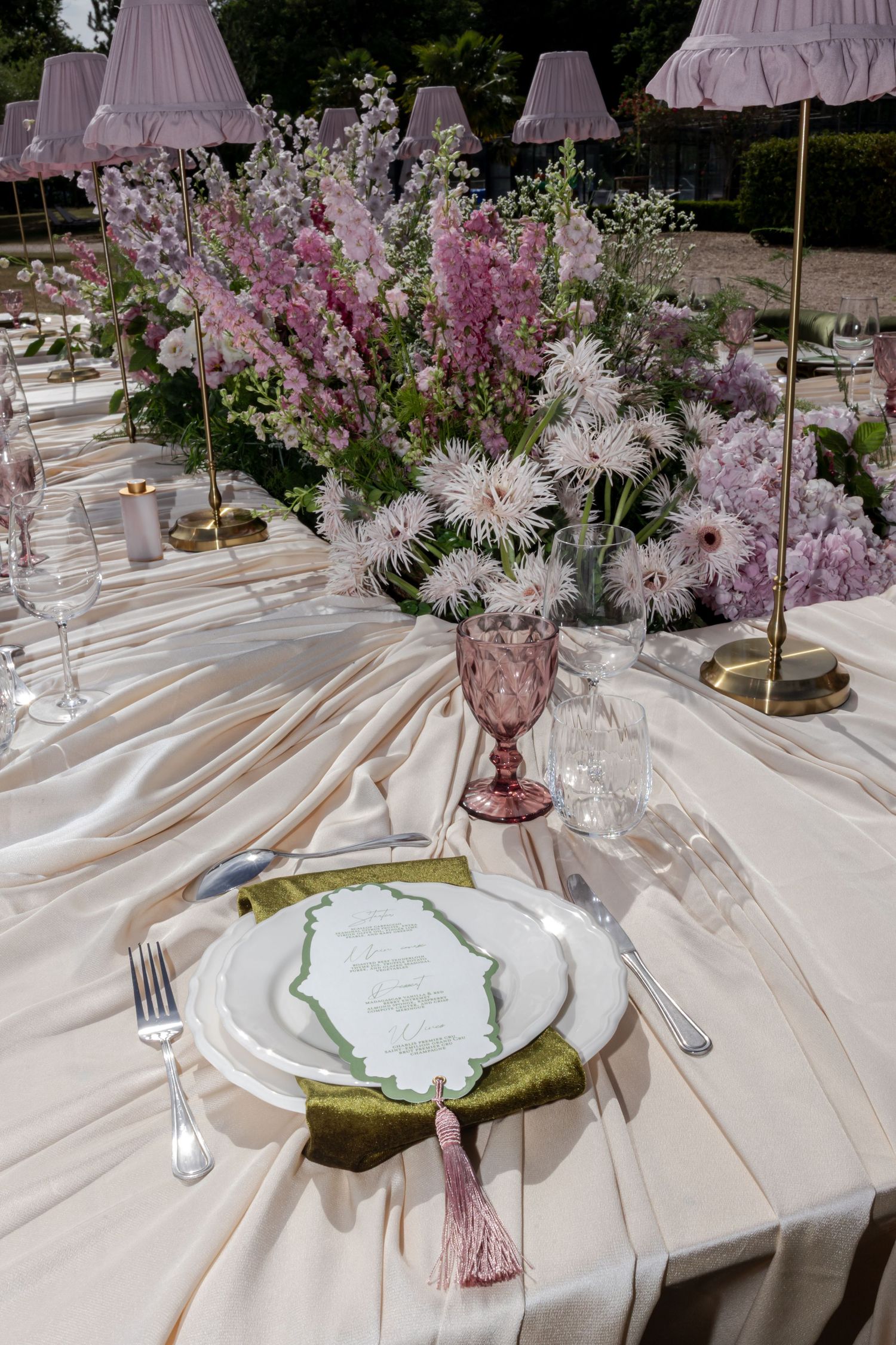 Elegant outdoor wedding table setting with pink floral centerpiece, draped ivory linen, green napkin, and pink goblet.