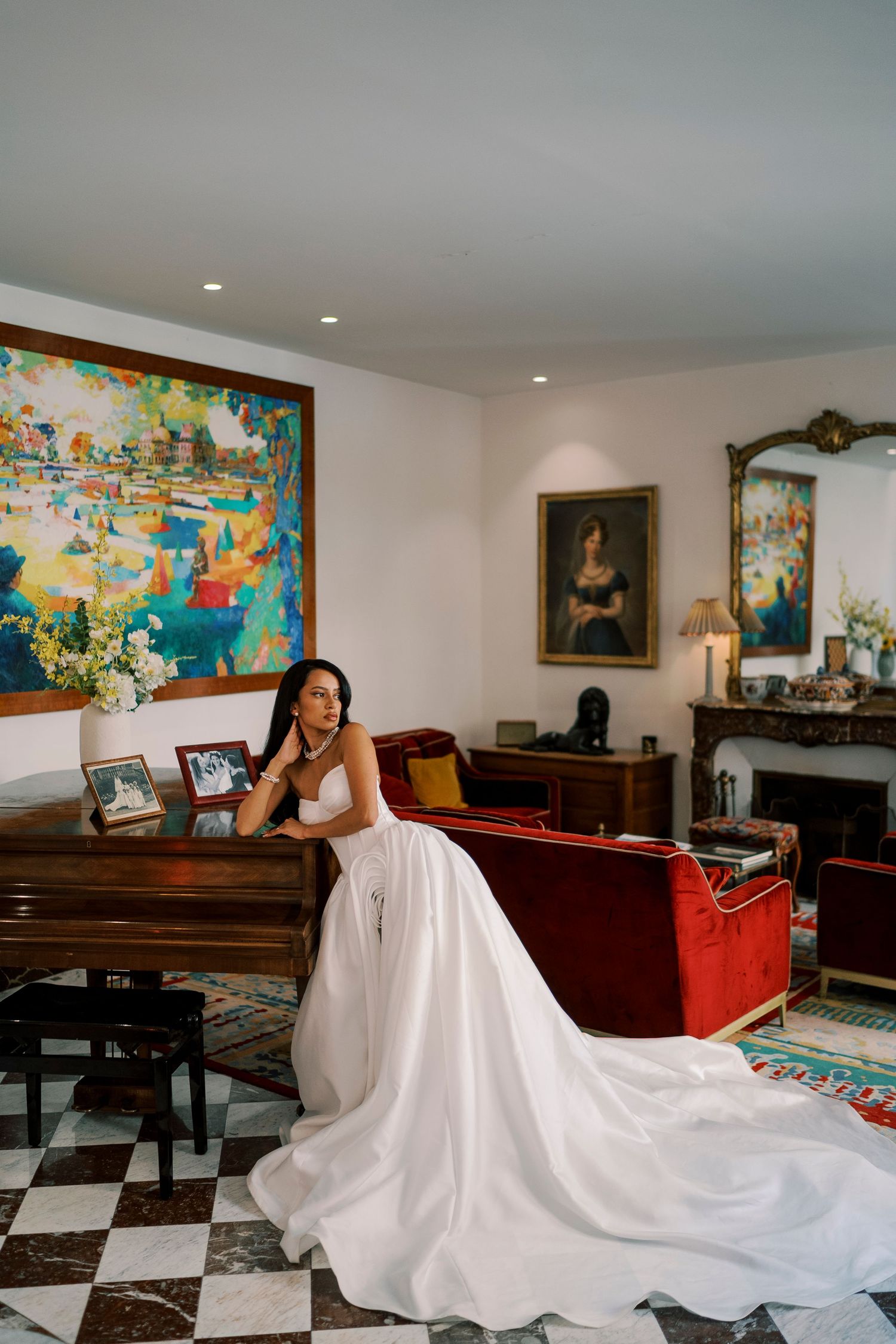 Bride in a white gown leans on a grand piano in an elegant room with colorful artwork and red velvet sofa.
