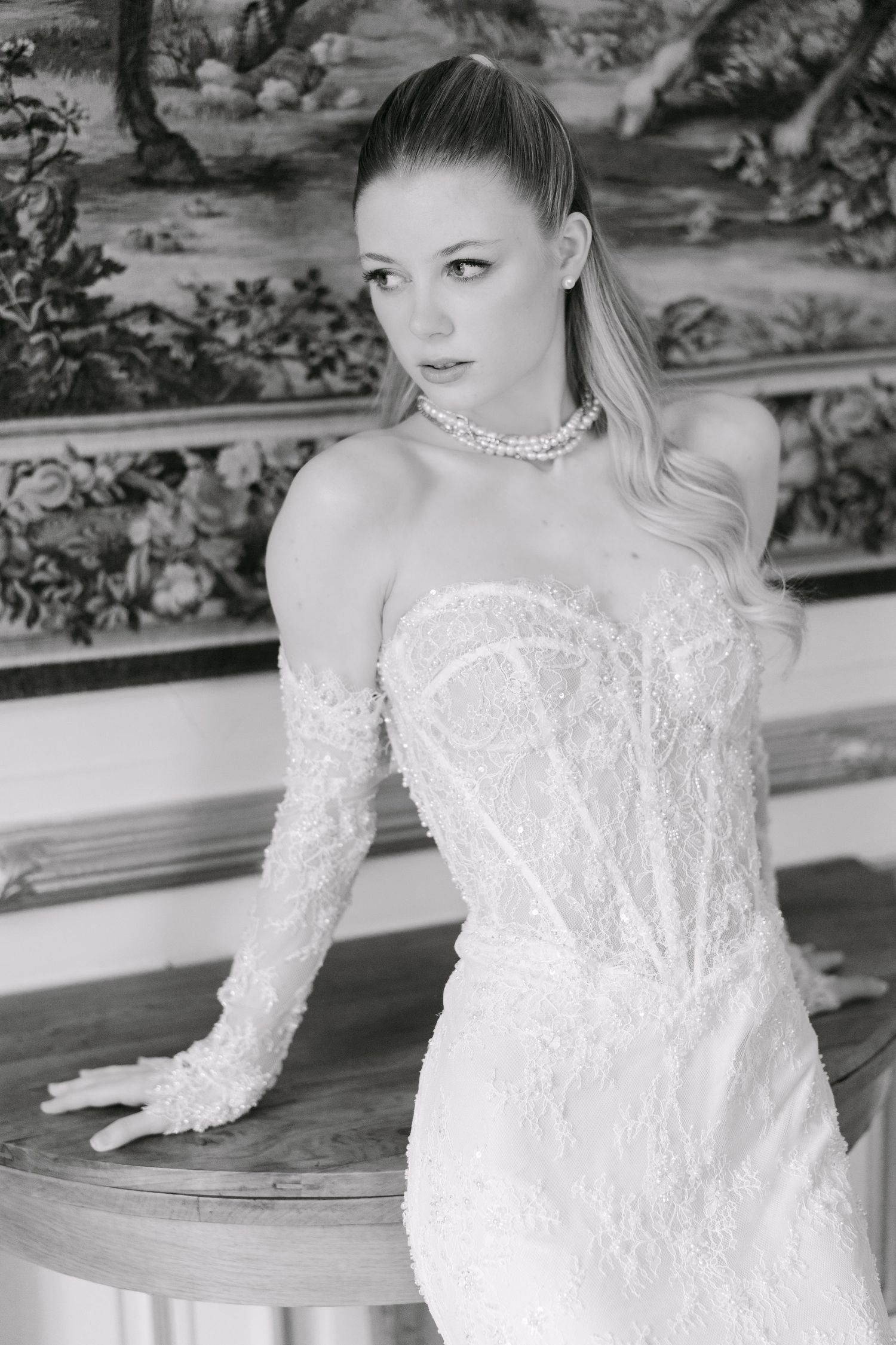Black and white photo of a bride in a strapless lace corset gown with long sleeves and pearl necklace.