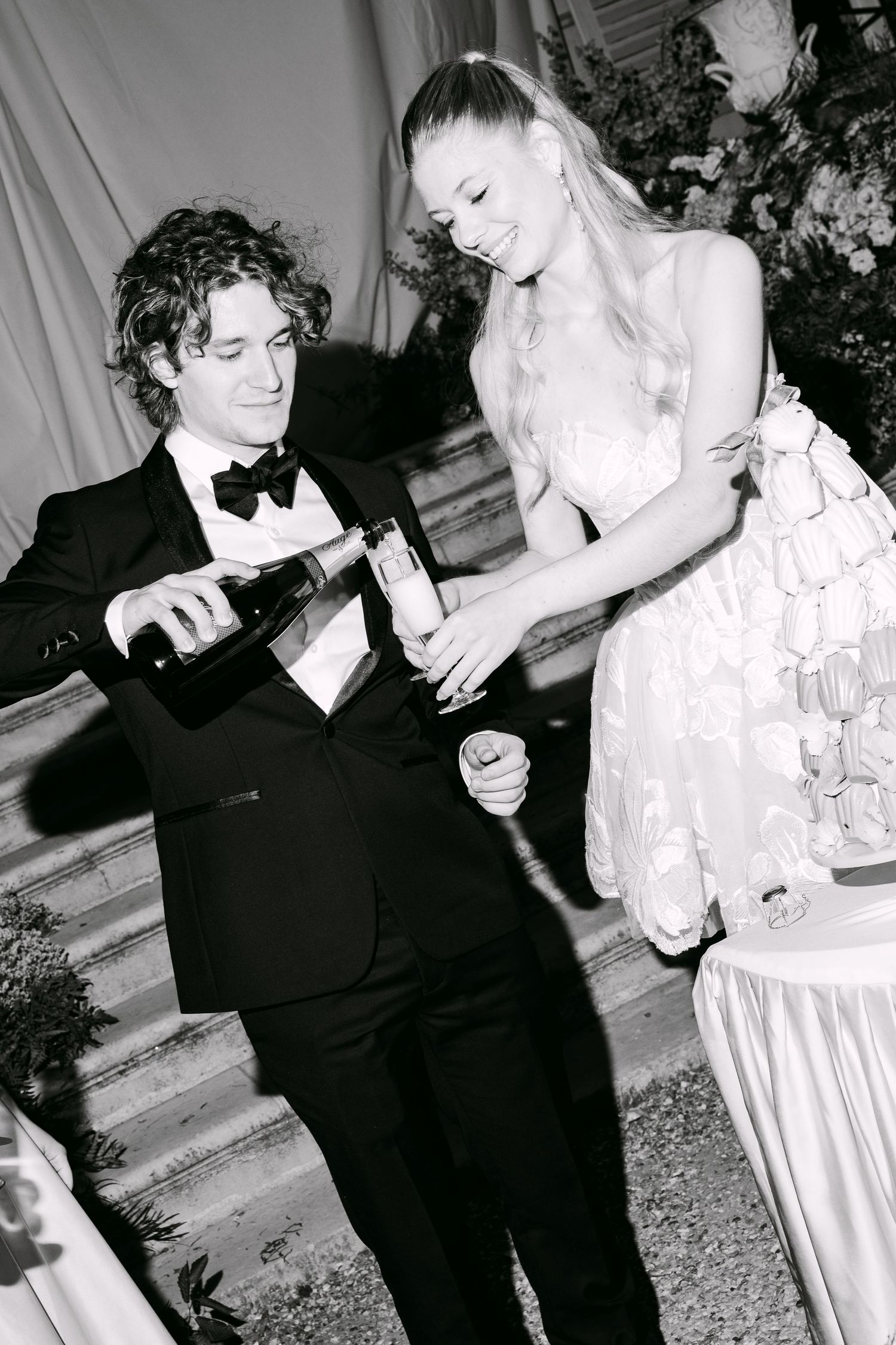 Black and white photo of a groom pouring champagne for a bride in a floral dress at a wedding reception.