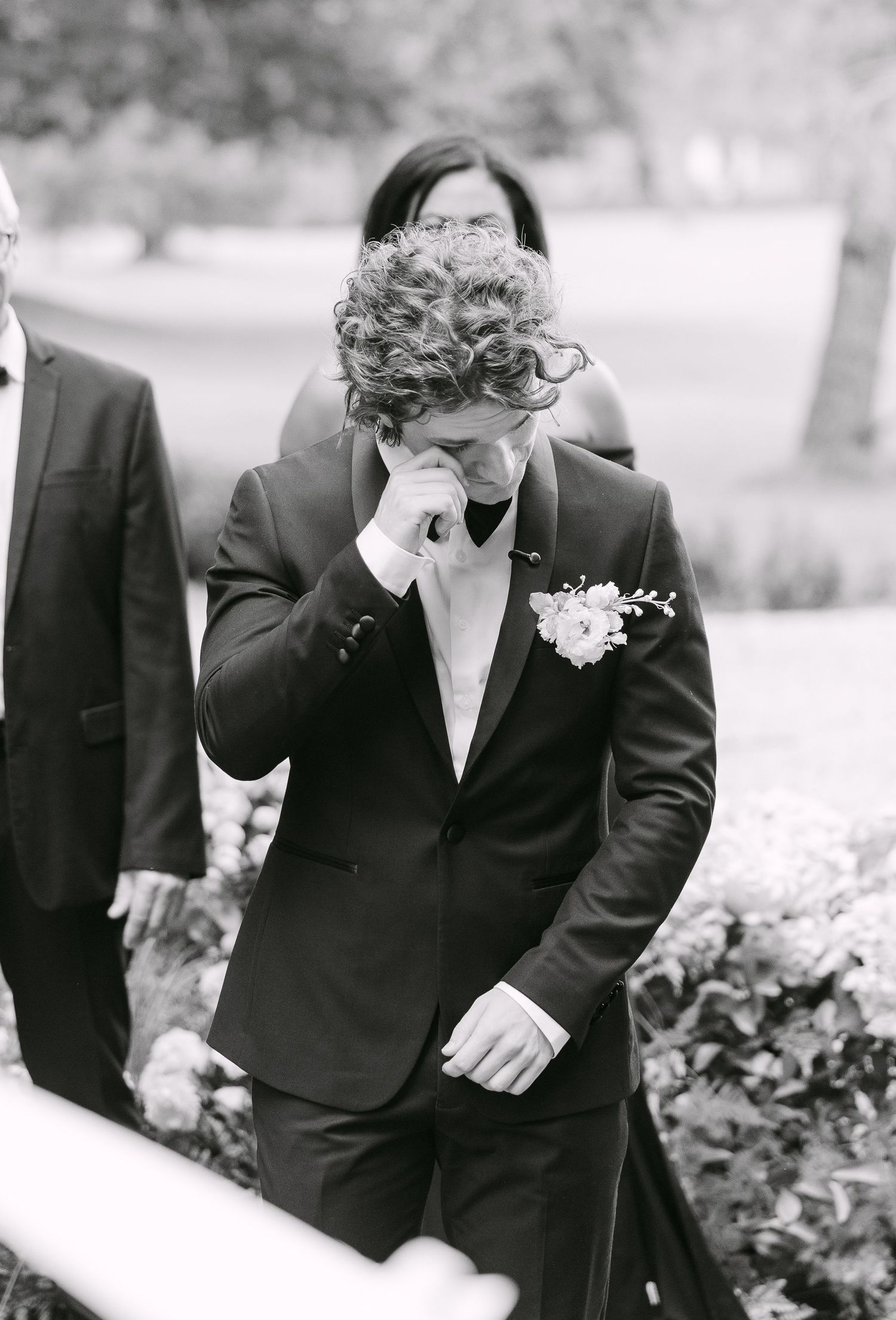 Emotional groom in black suit with boutonniere wiping tears during outdoor wedding ceremony, black and white photo.