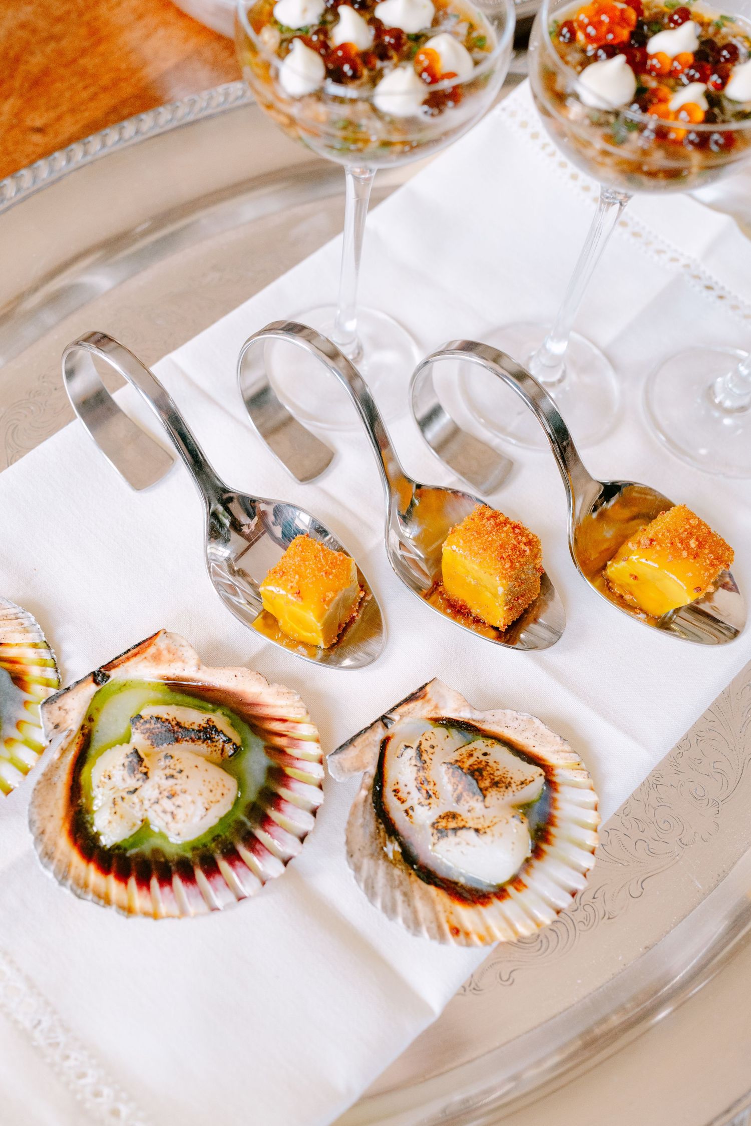 Elegant wedding canapés featuring scallops in shells and golden bites on silver spoons on a silver tray.