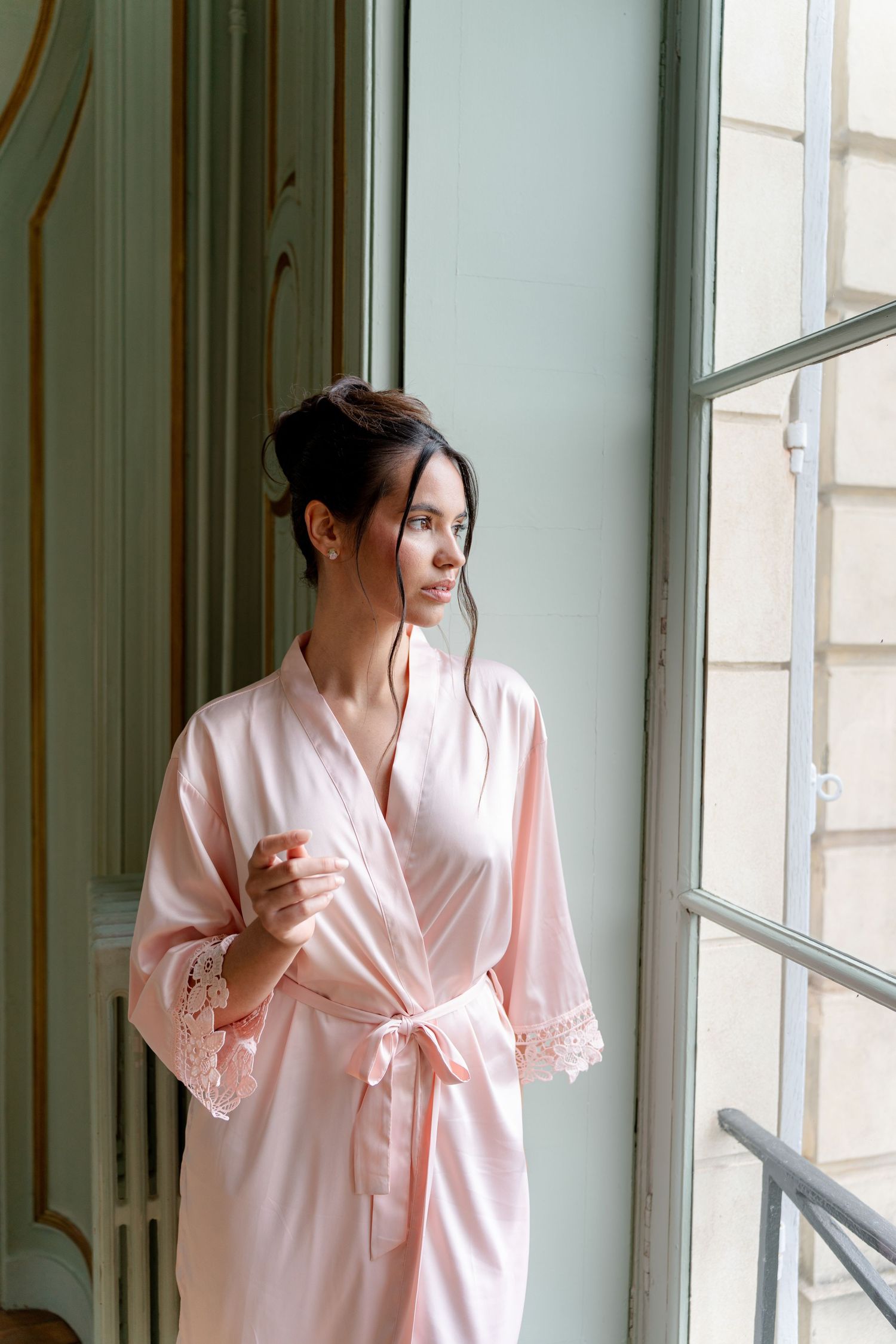 Woman in pink satin robe with lace trim standing by a large window in an elegant room.