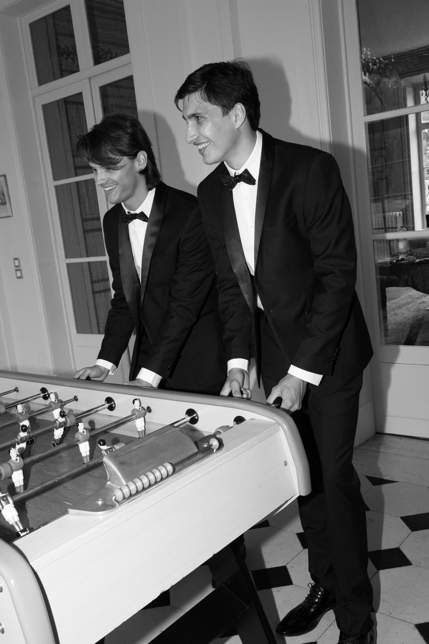 Two men in tuxedos playing foosball at an elegant event, black and white photo.