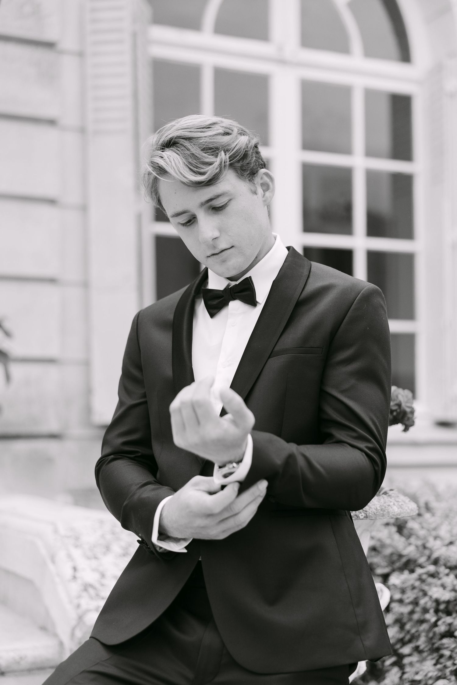 Young man in black tuxedo with bow tie adjusting cufflinks outdoors in elegant black and white photo.