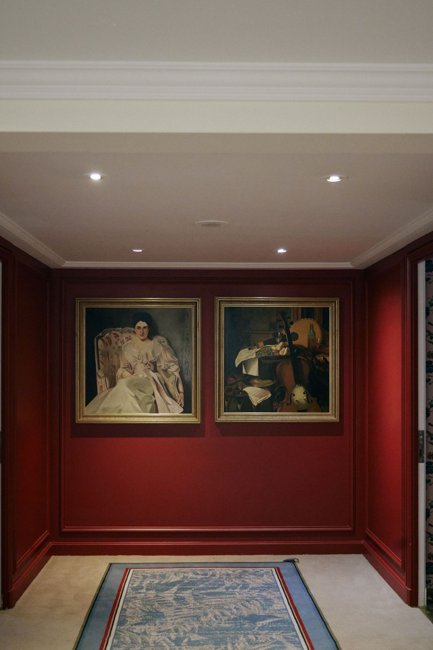 Elegant red-walled hallway featuring two gold-framed classical paintings, recessed lighting, and a decorative blue area rug.