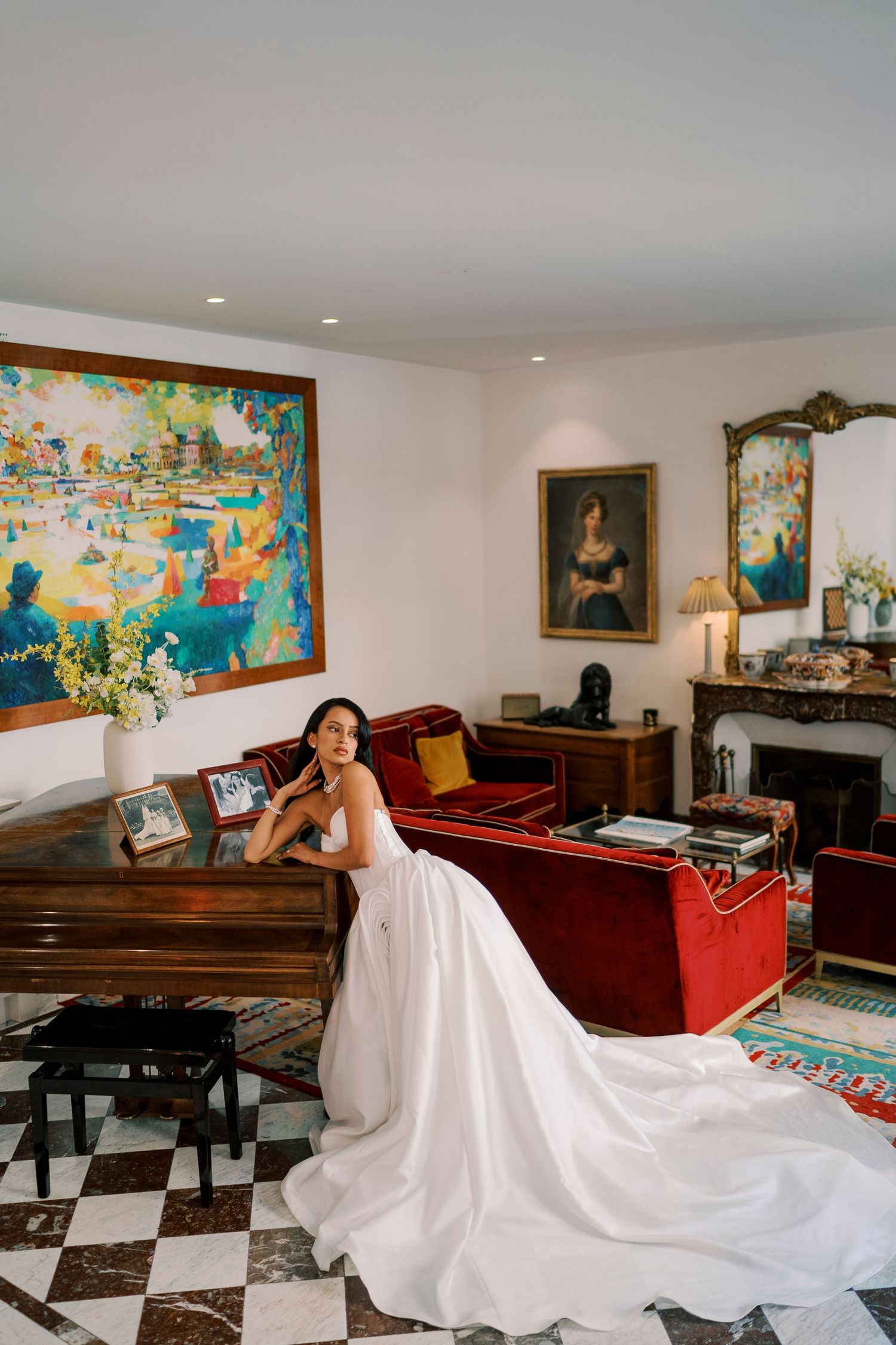Bride in white ball gown wedding dress posing in elegant room with colorful artwork and red velvet sofa.