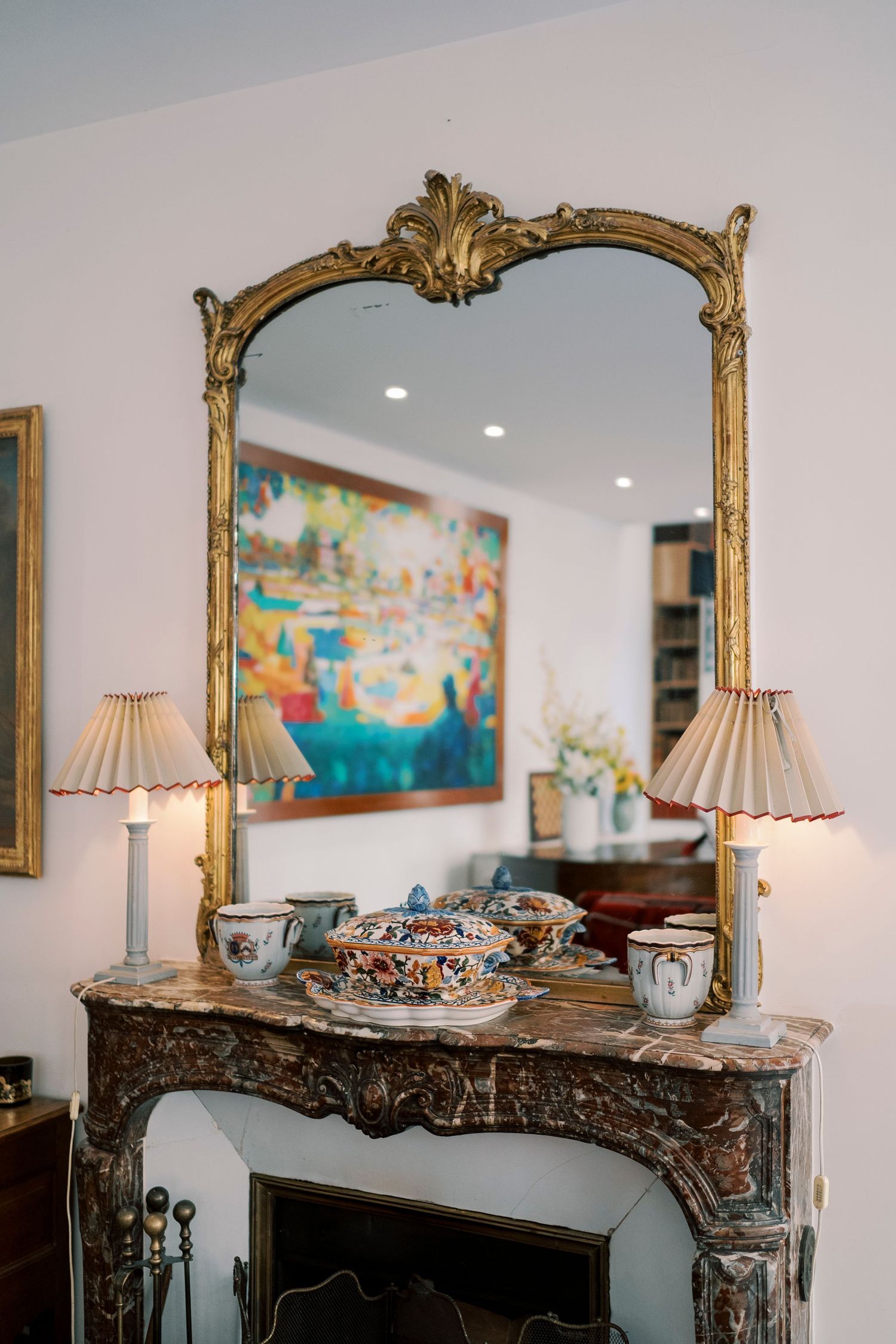 Ornate gold-framed mirror above marble fireplace mantel with decorative porcelain dishes and matching table lamps.