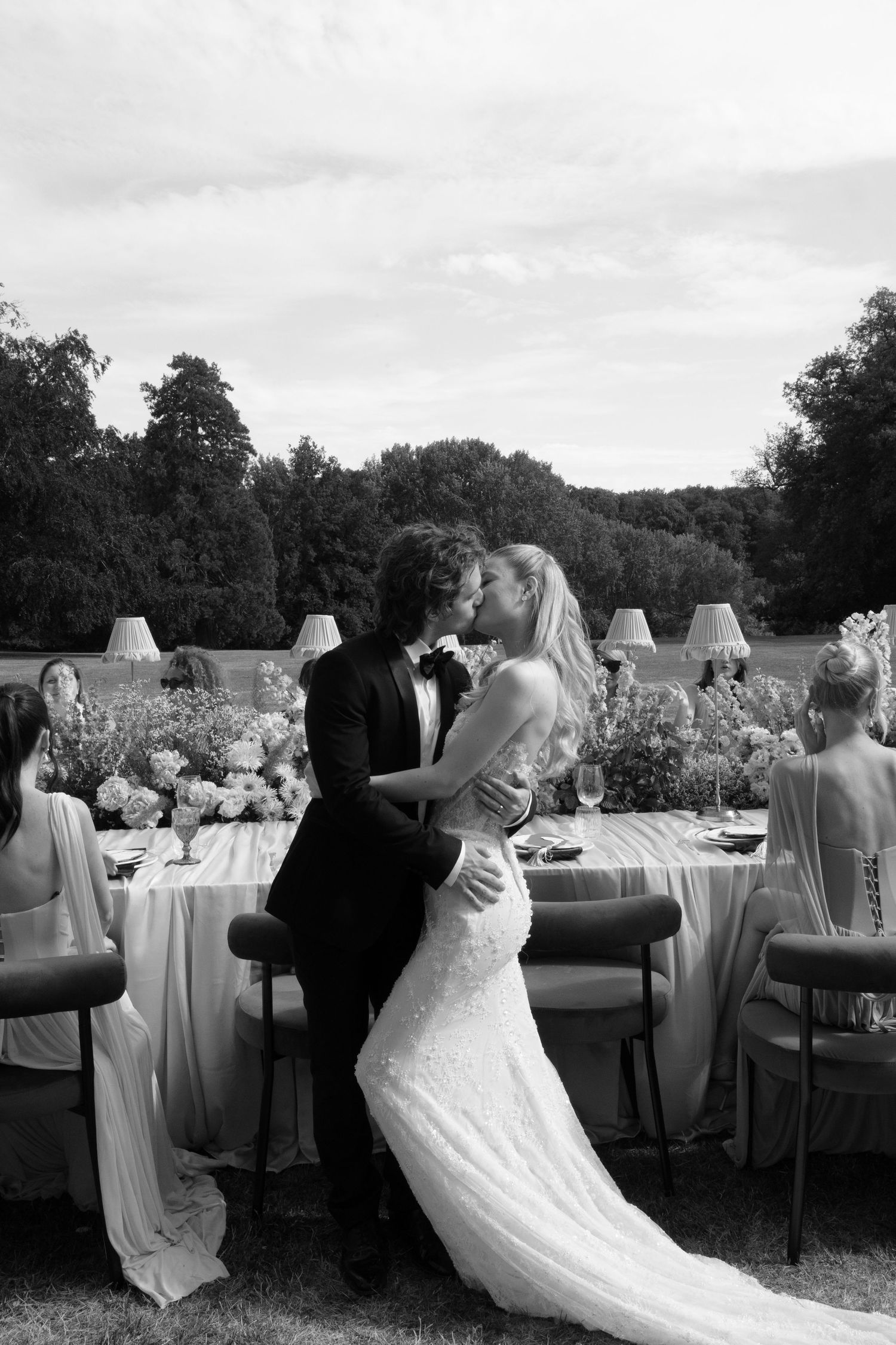 Black and white photo of a bride and groom sharing a kiss at an outdoor wedding reception dinner table.