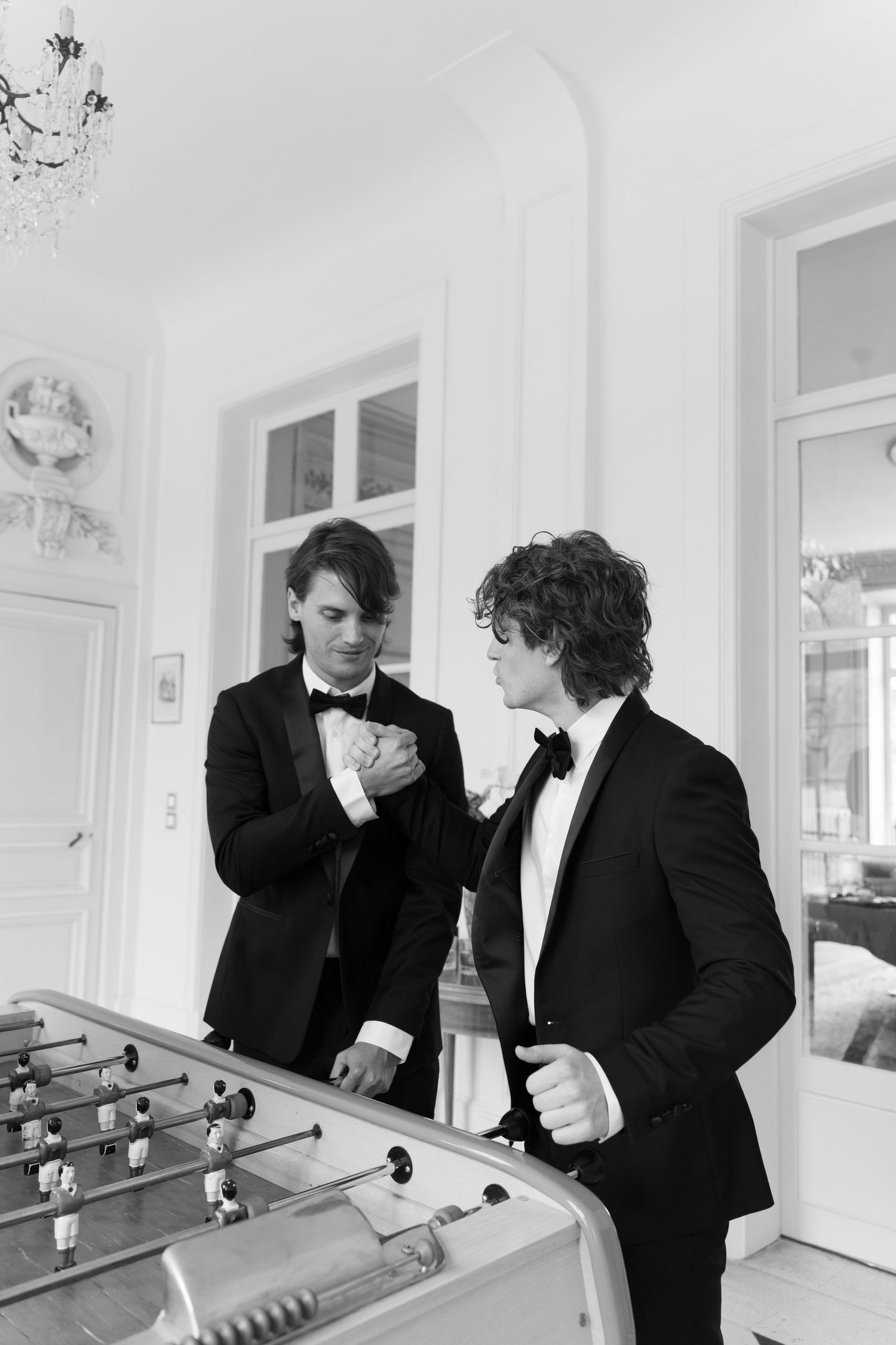 Two men in tuxedos with bow ties stand by a foosball table in an elegant white room, black and white photo.