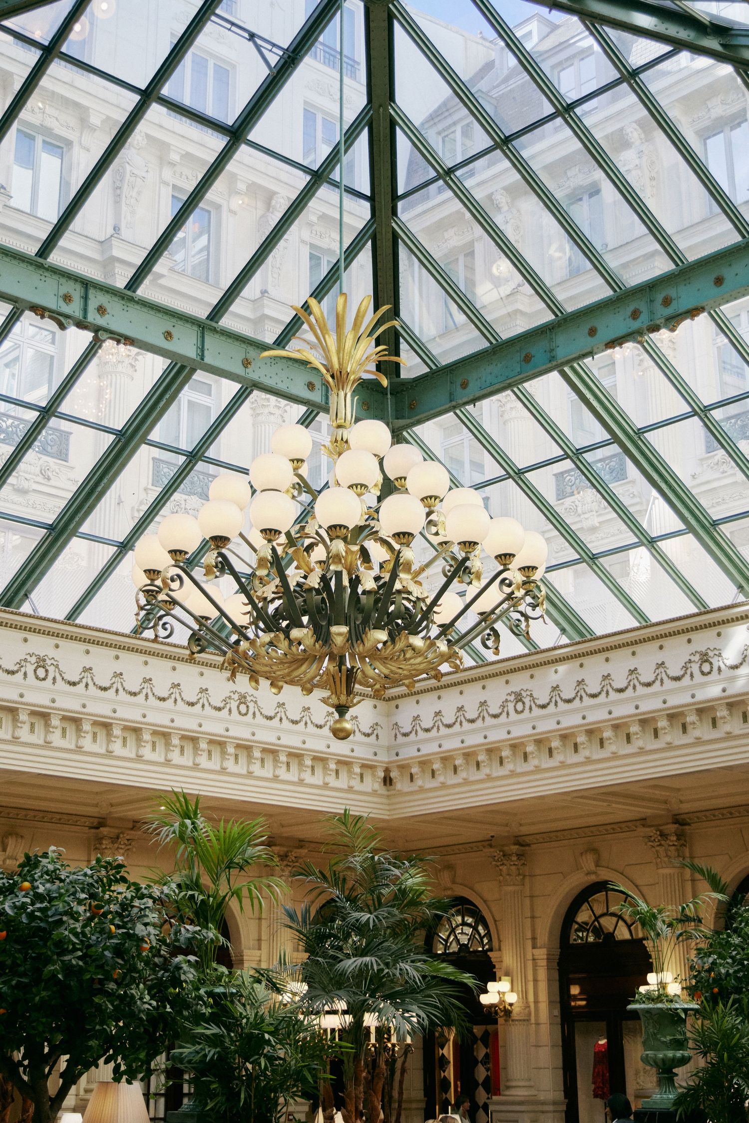 Elegant chandelier hanging beneath a glass pyramid ceiling in a grand classical interior with palm trees.