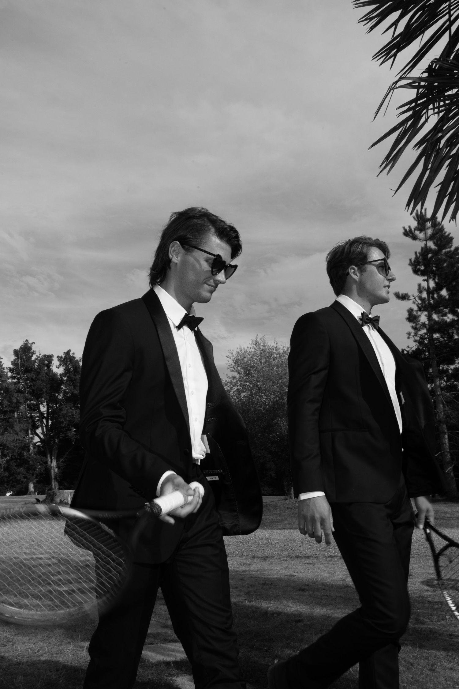 Two men in black tuxedos and sunglasses walk outdoors near palm trees in a stylish black and white photo.