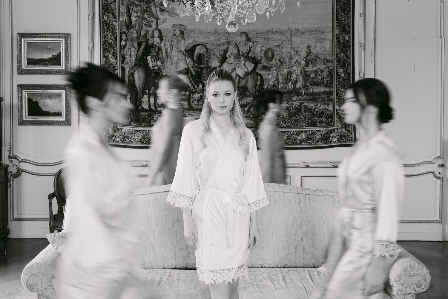 Black and white photo of a bride in a white robe standing still while attendants move around her in a elegant room.