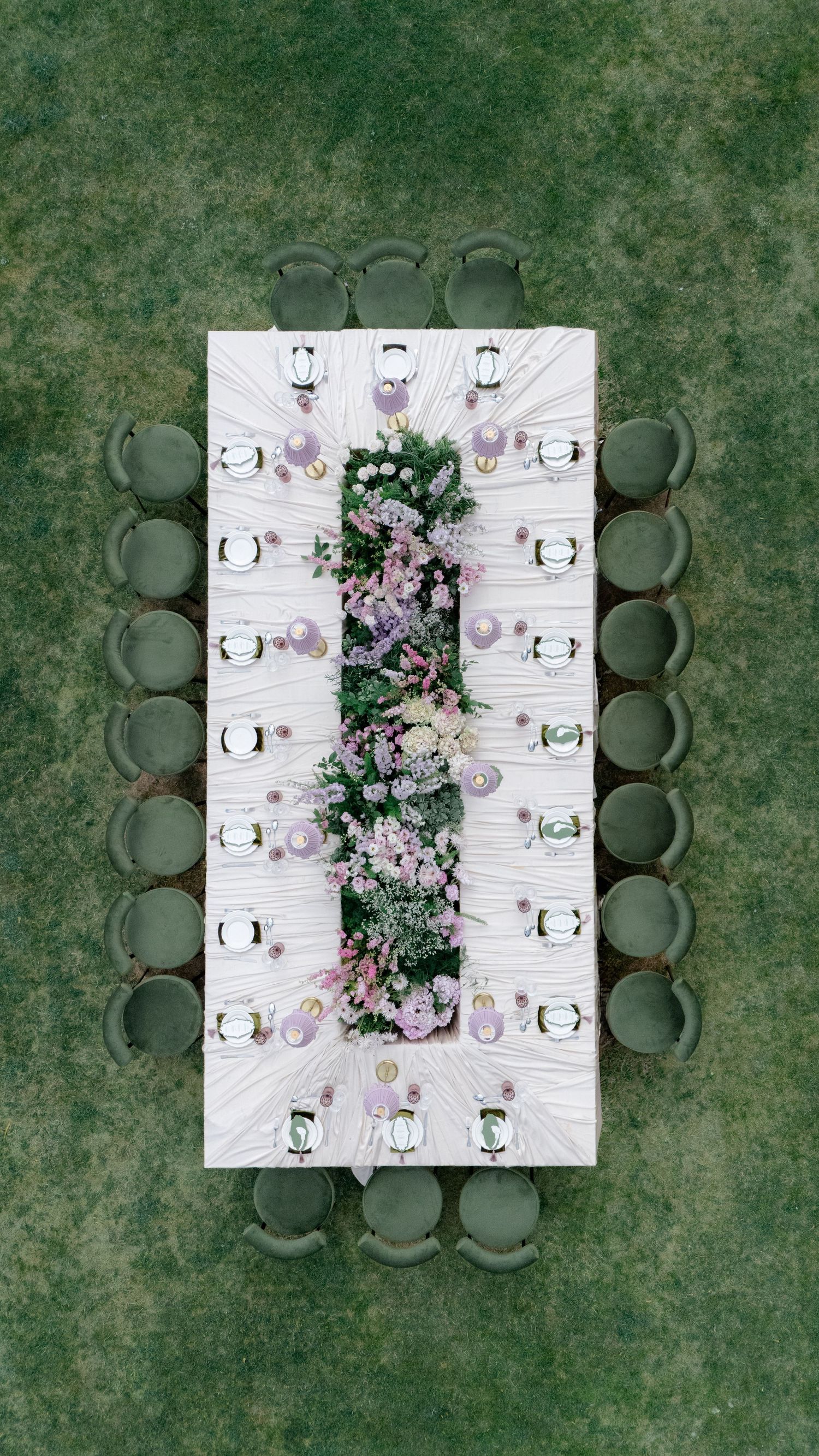 Aerial view of an elegant outdoor wedding reception table with lush floral centerpiece, purple accents, and green chairs on grass.