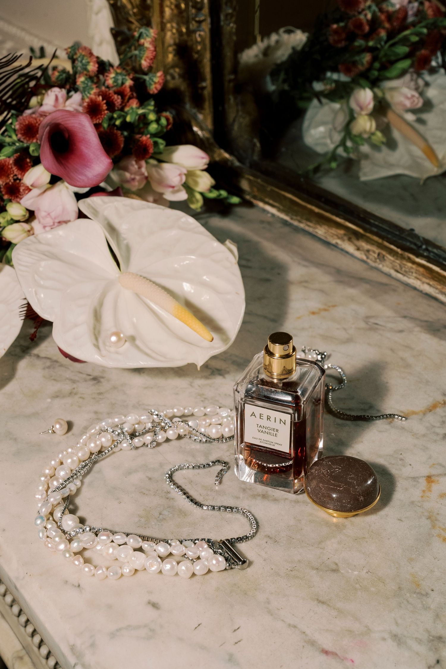 AERIN perfume bottle with pearl necklace, white anthurium flower, and floral bouquet on marble surface near mirror.