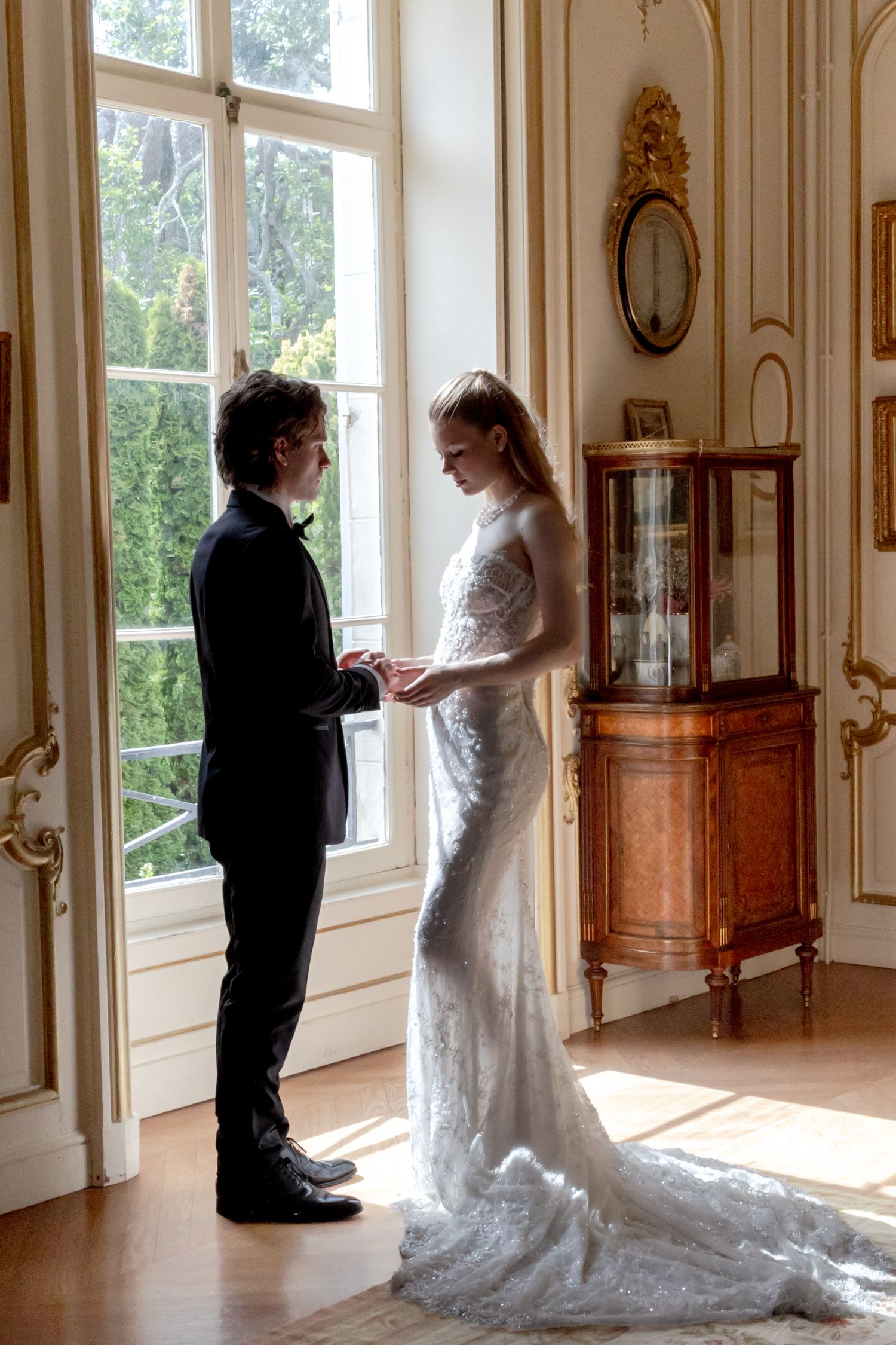 Bride and groom exchange rings in an elegant gold-adorned room with large windows and antique furniture.
