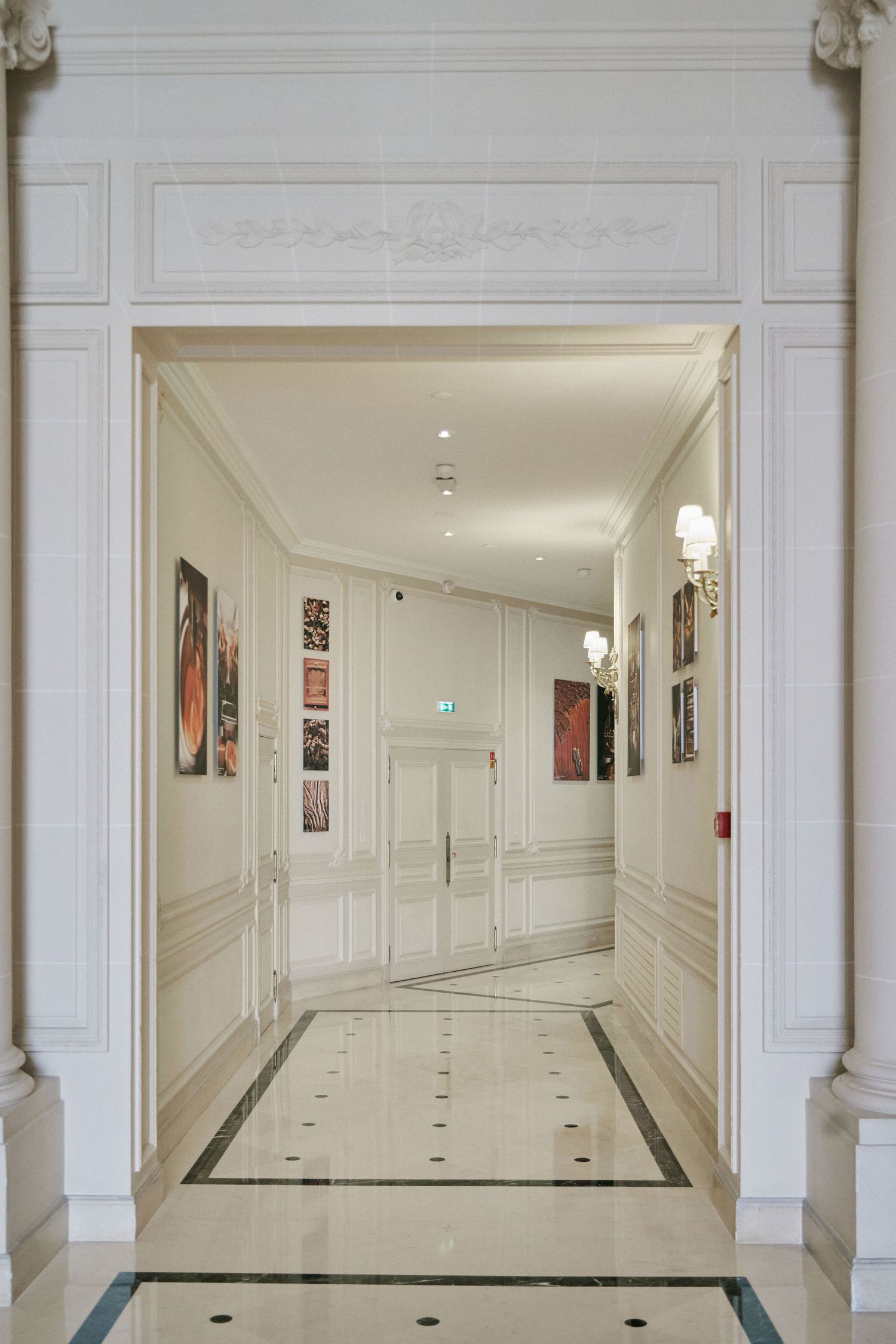 Elegant hotel corridor with white wainscoting, marble tile floors, wall art, and ornate doorway leading to double doors.