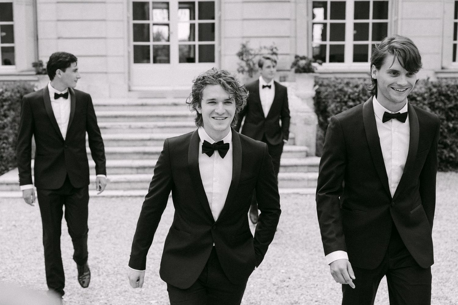 Groomsmen in black tuxedos smile and walk outside an elegant French chateau in black and white photo.
