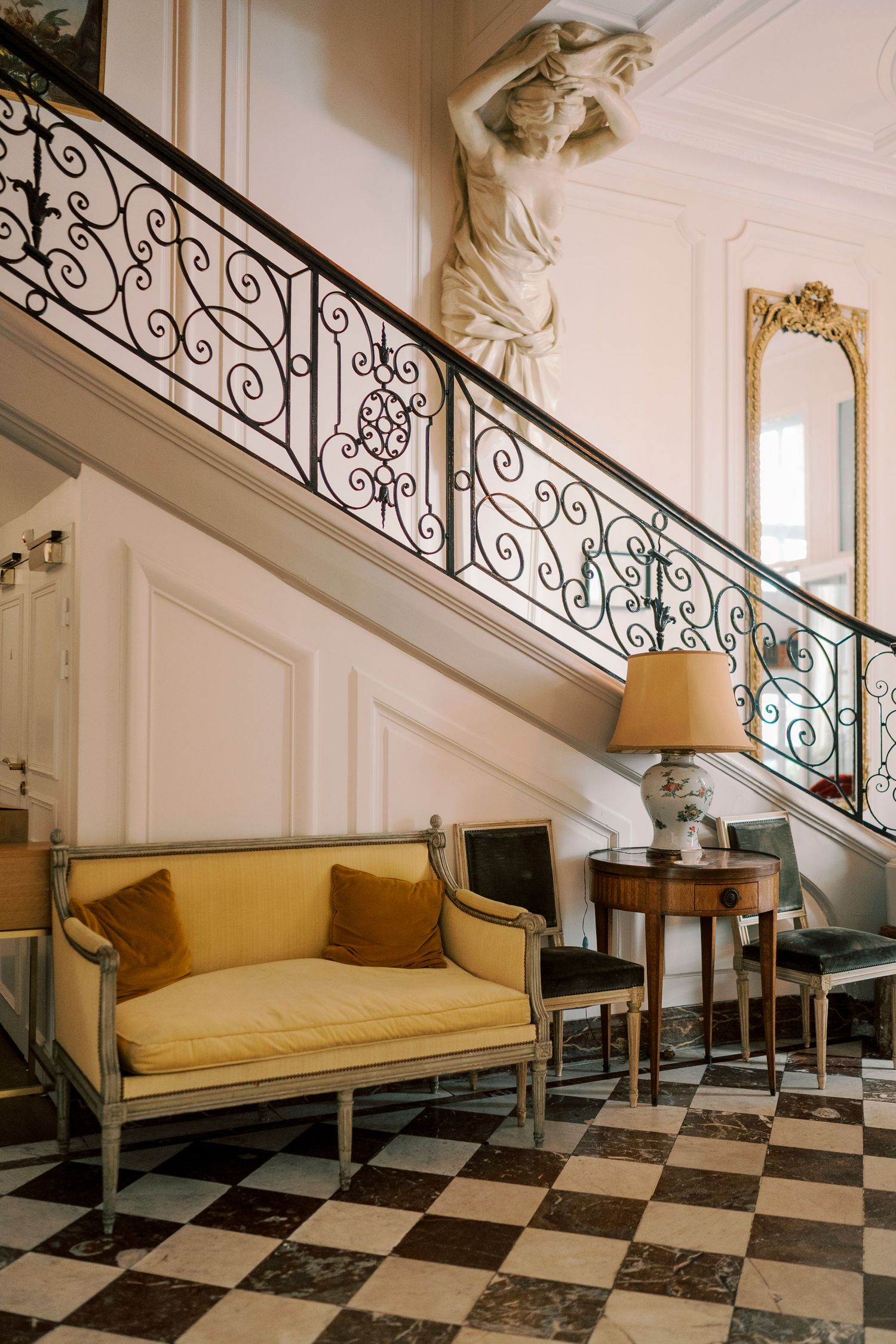 Elegant French foyer with ornate wrought iron staircase, yellow velvet settee, checkered marble floor, and classical sculpture.
