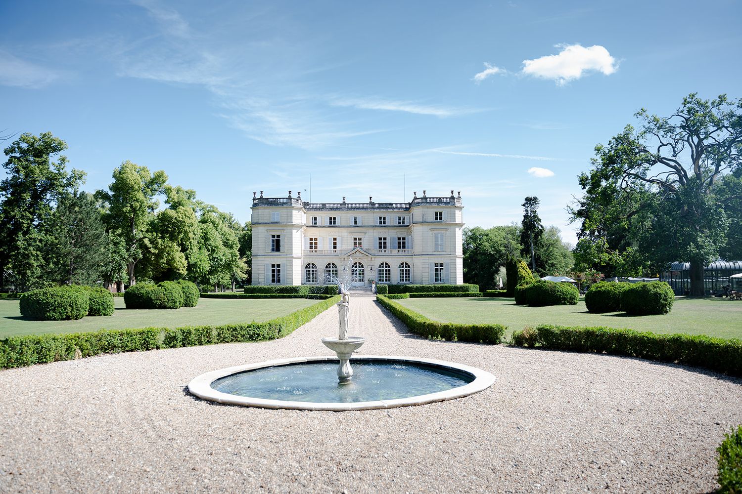Elegant French chateau with circular fountain, manicured hedges, and gravel driveway under blue sky.