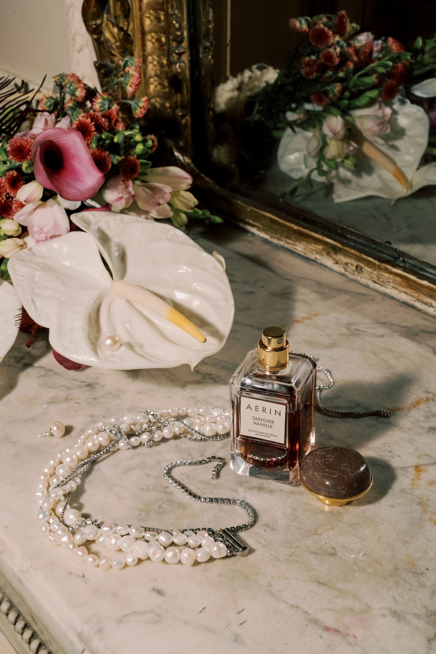 Elegant vanity scene with AERIN perfume, pearl necklace, compact mirror, and floral bouquet on marble surface.