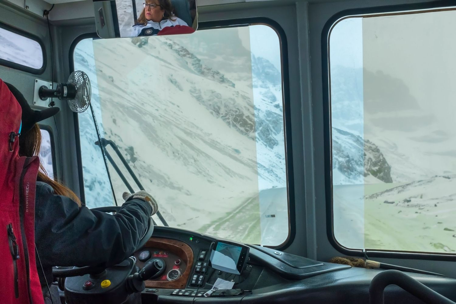 Driver navigating a mountain bus through snowy alpine terrain with panoramic windshield views of glaciers.