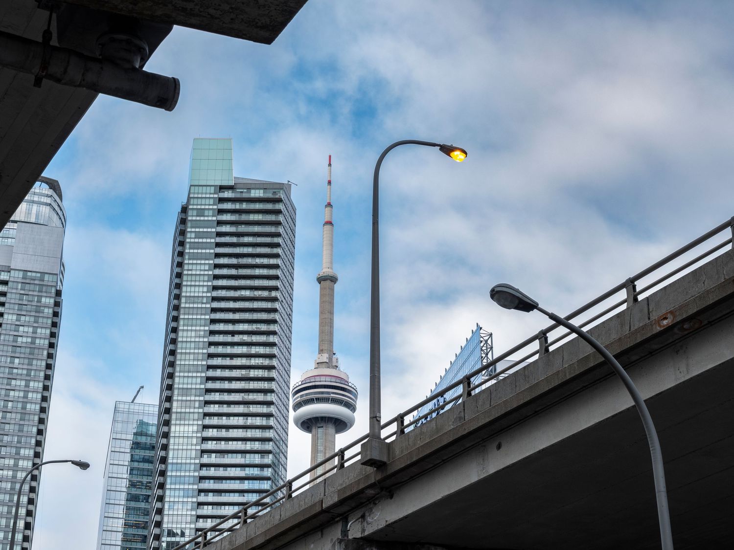 Toronto skyline featuring the iconic CN Tower, modern skyscrapers, an elevated highway, and street lamps under a partly cloudy sky.