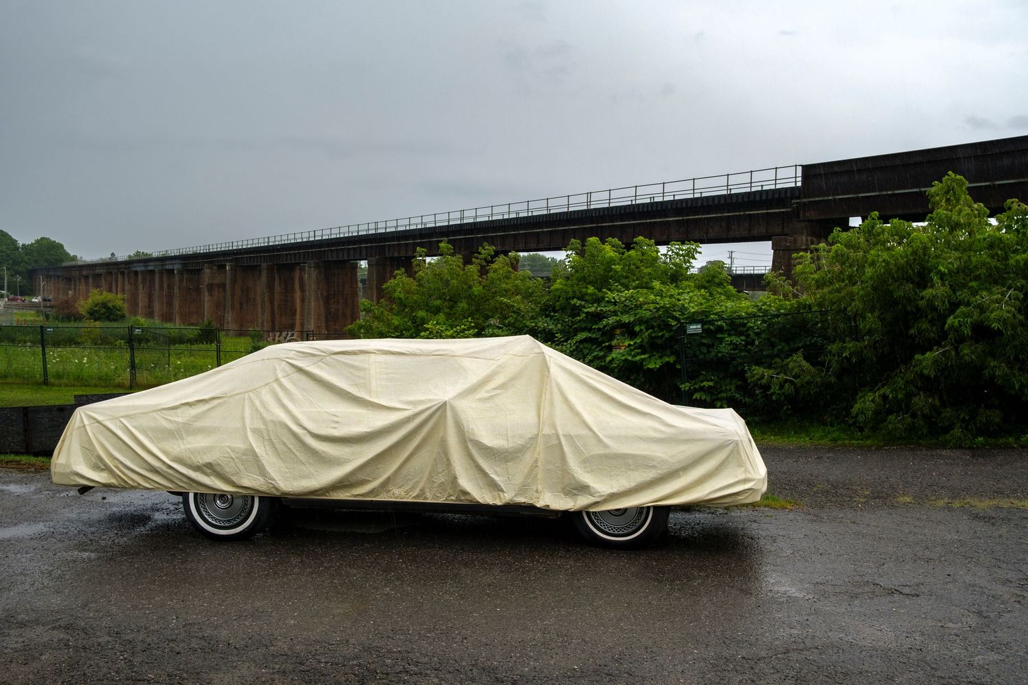 Classic car covered with a beige protective cover parked on wet pavement near a bridge on a rainy day.