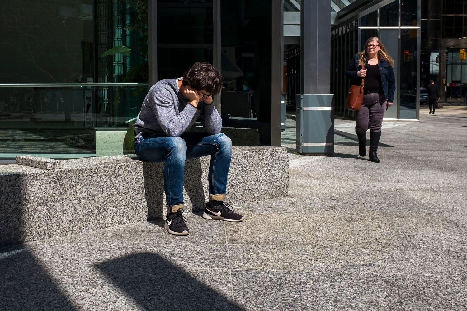 A distressed man sits hunched over on a granite bench outside a modern building in an urban setting.