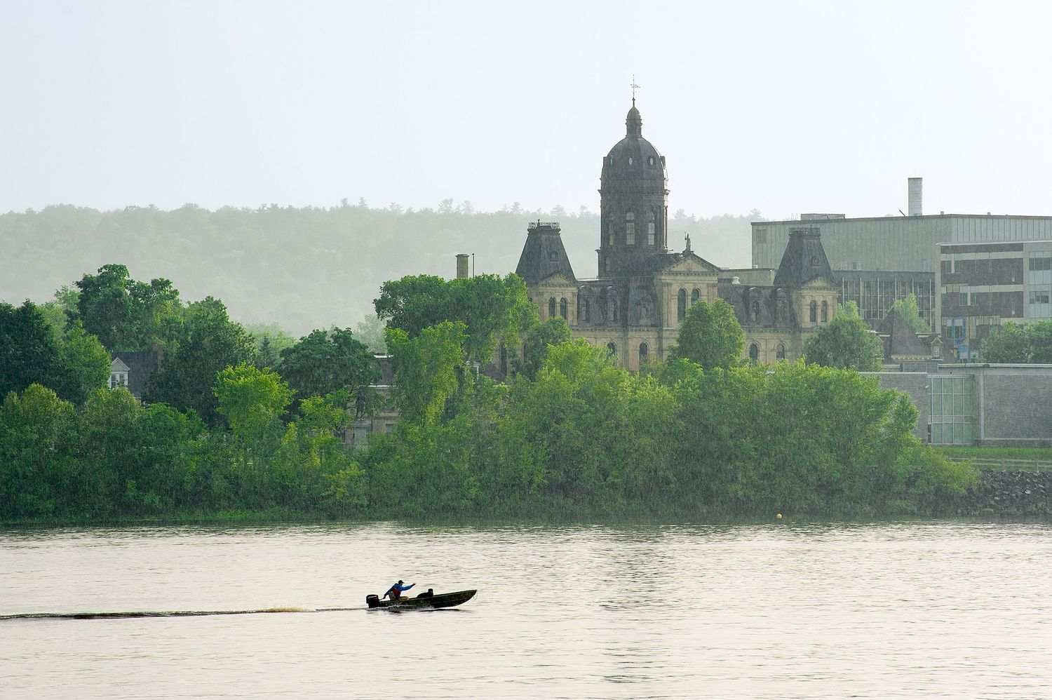 A small boat glides along a calm river with lush green trees and a historic church building visible in the misty background.