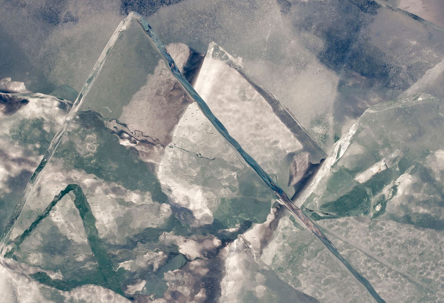 Aerial view of cracked ice sheets forming geometric patterns on a frozen lake surface with blue-green hues.