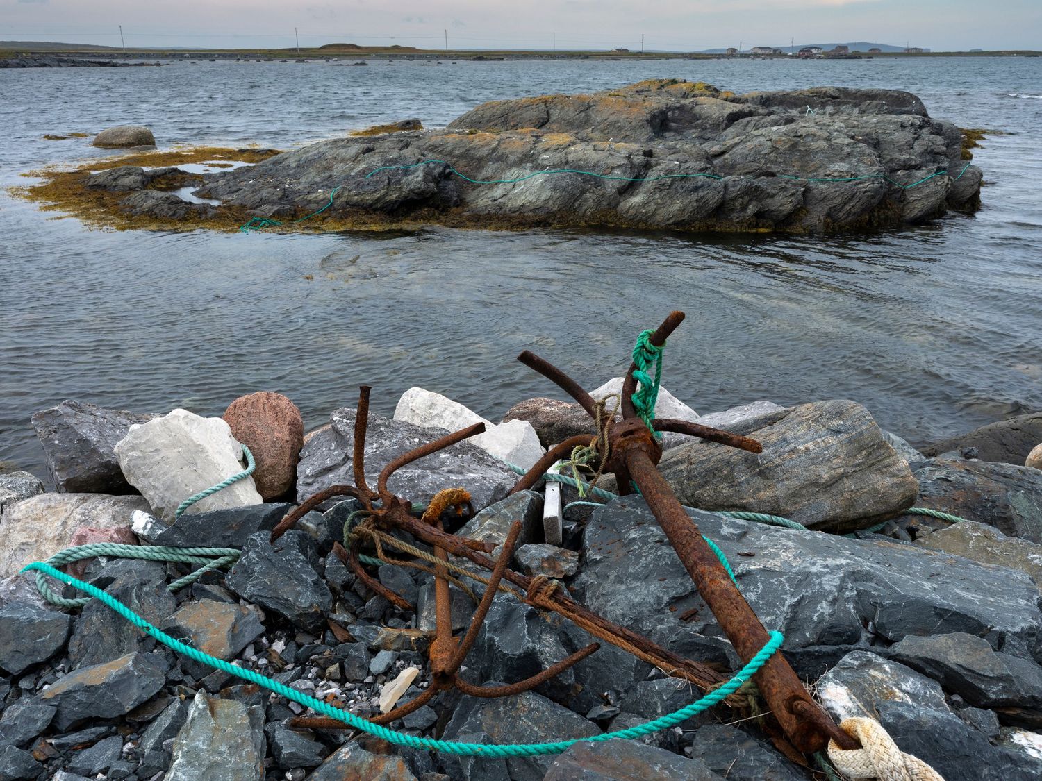 Rusty anchor with teal rope on rocky shoreline, with ocean water and large rocks in background.