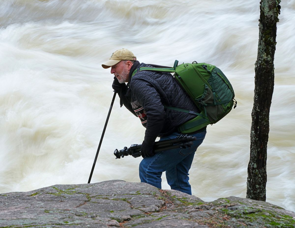 Hiker with green backpack and trekking pole navigating rocky terrain near rushing rapids in a forest.