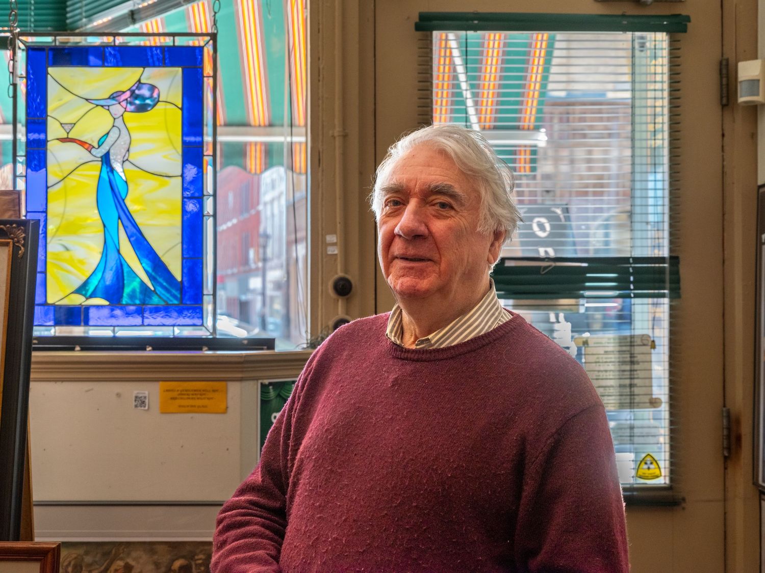 Elderly man in maroon sweater stands near colorful stained glass artwork in a shop.