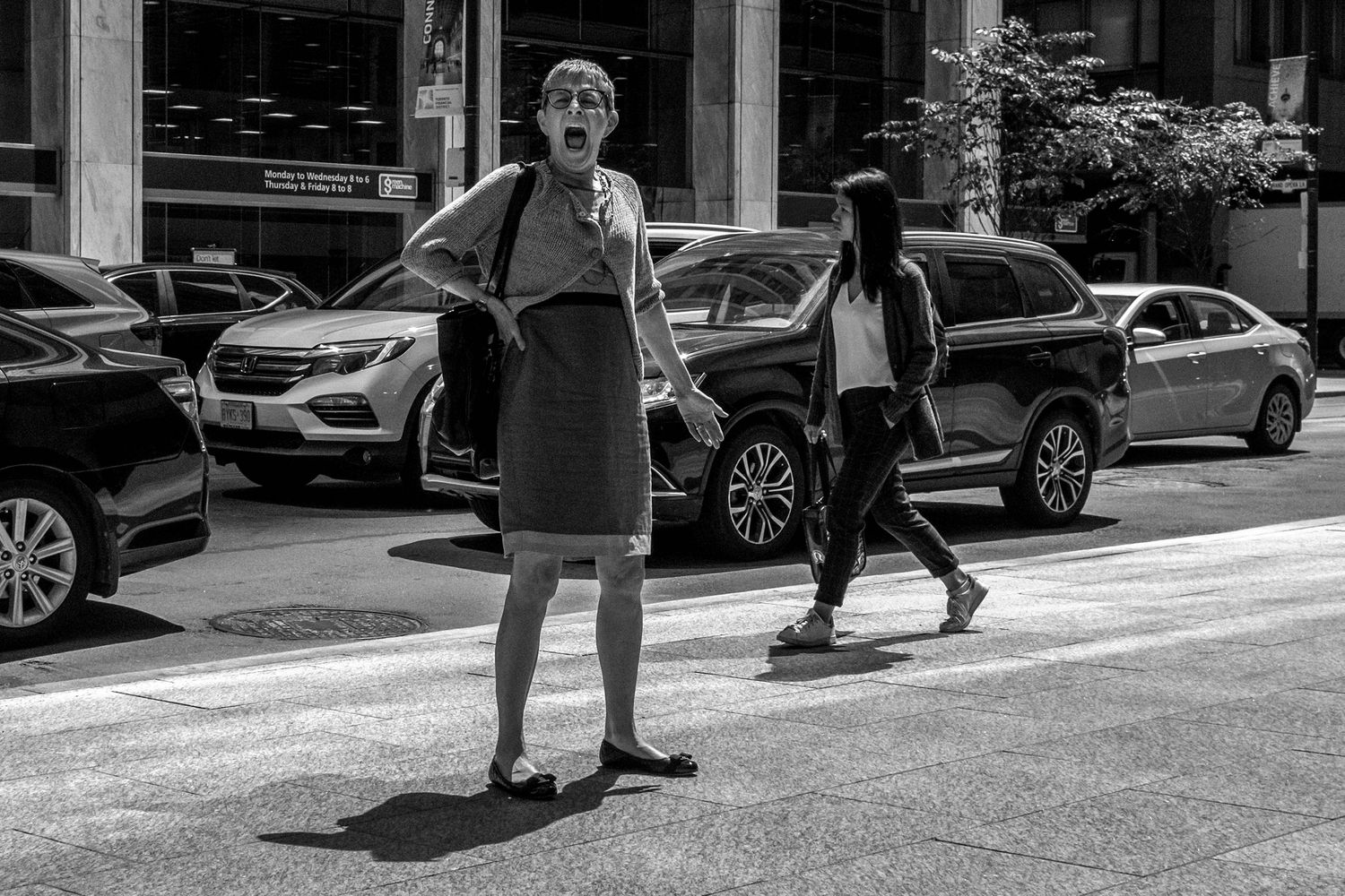 Black and white street photo of a woman posing on a crosswalk with cars and a pedestrian in the background.