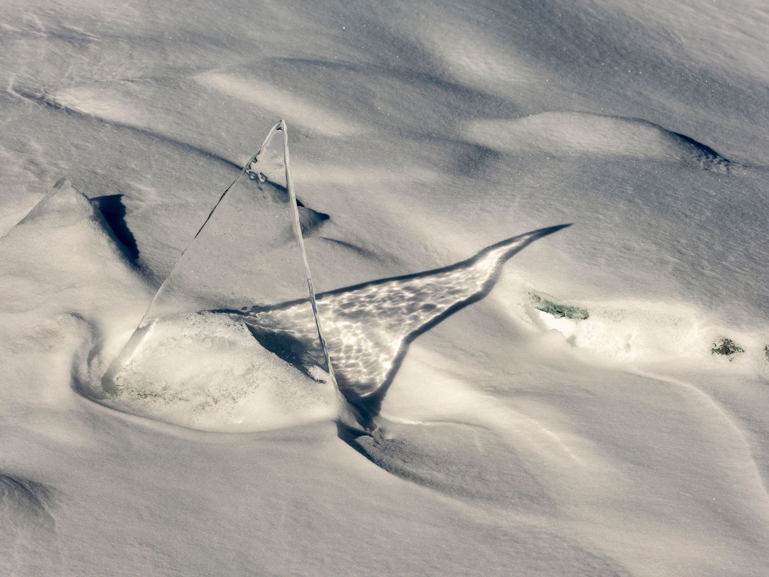 Aerial view of snow-covered landscape with wind-sculpted drifts and a small marker pole casting shadows on white terrain.