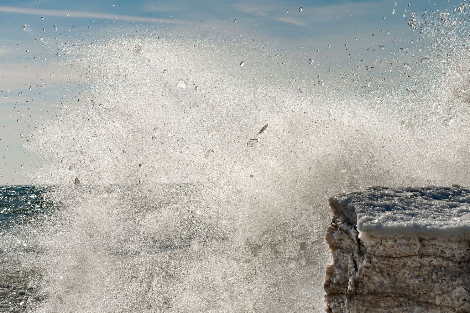 Powerful ocean wave crashing against rocky shoreline, sending white sea spray high into the air under a blue sky.