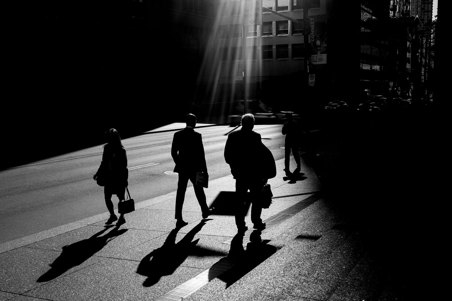 Black and white photo of silhouetted pedestrians crossing a sunlit urban street with dramatic shadows.