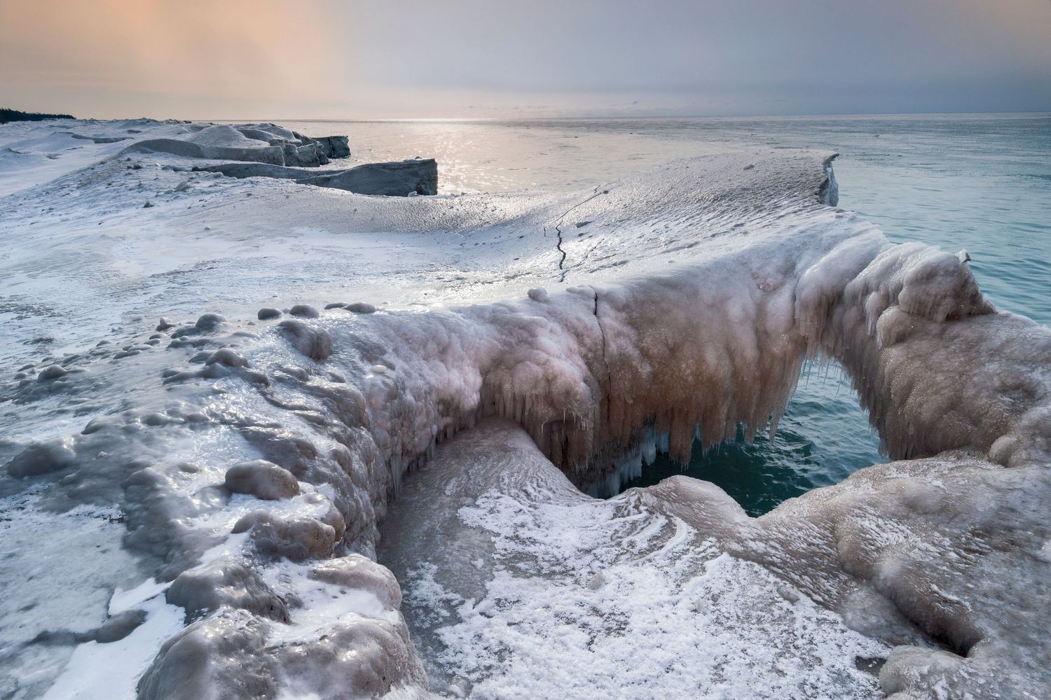 Ice-covered rocky shoreline with frozen formations over turquoise water under a misty winter sky.