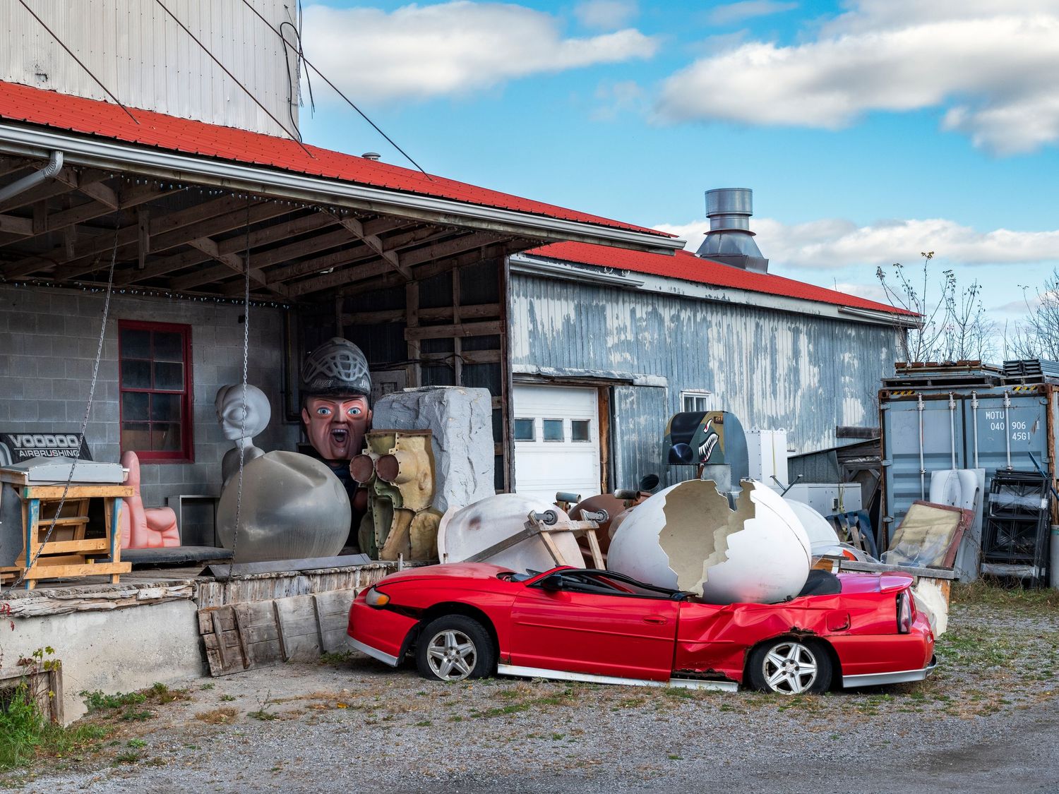 Man peering over giant egg sculptures beside a red convertible at a quirky rural junkyard art installation.
