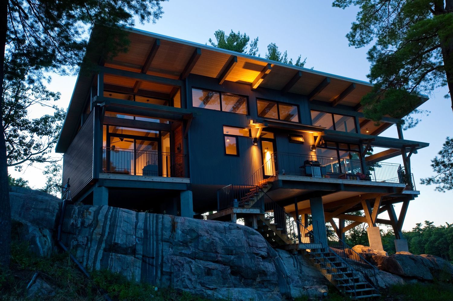 Modern two-story cabin on rocky hillside glows warmly at dusk, featuring large windows, wood beams, and elevated deck among trees.