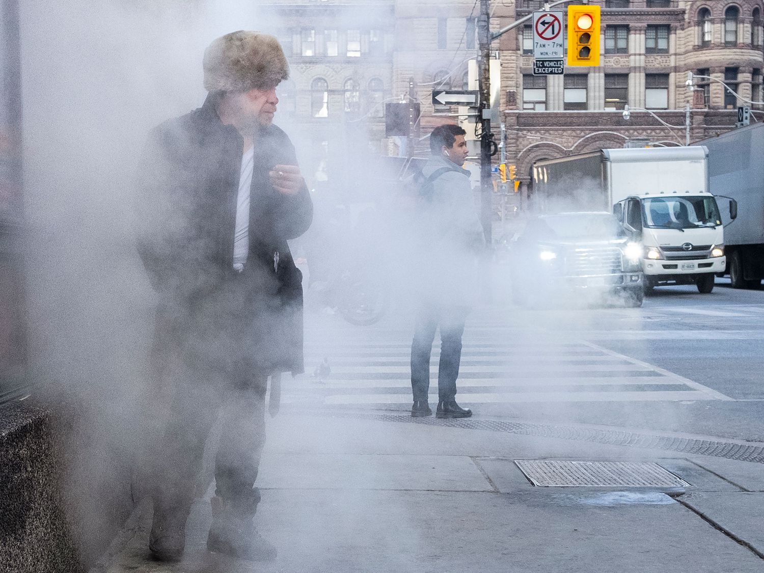 Two people standing on a misty urban sidewalk surrounded by steam, with city traffic and buildings visible in the background.