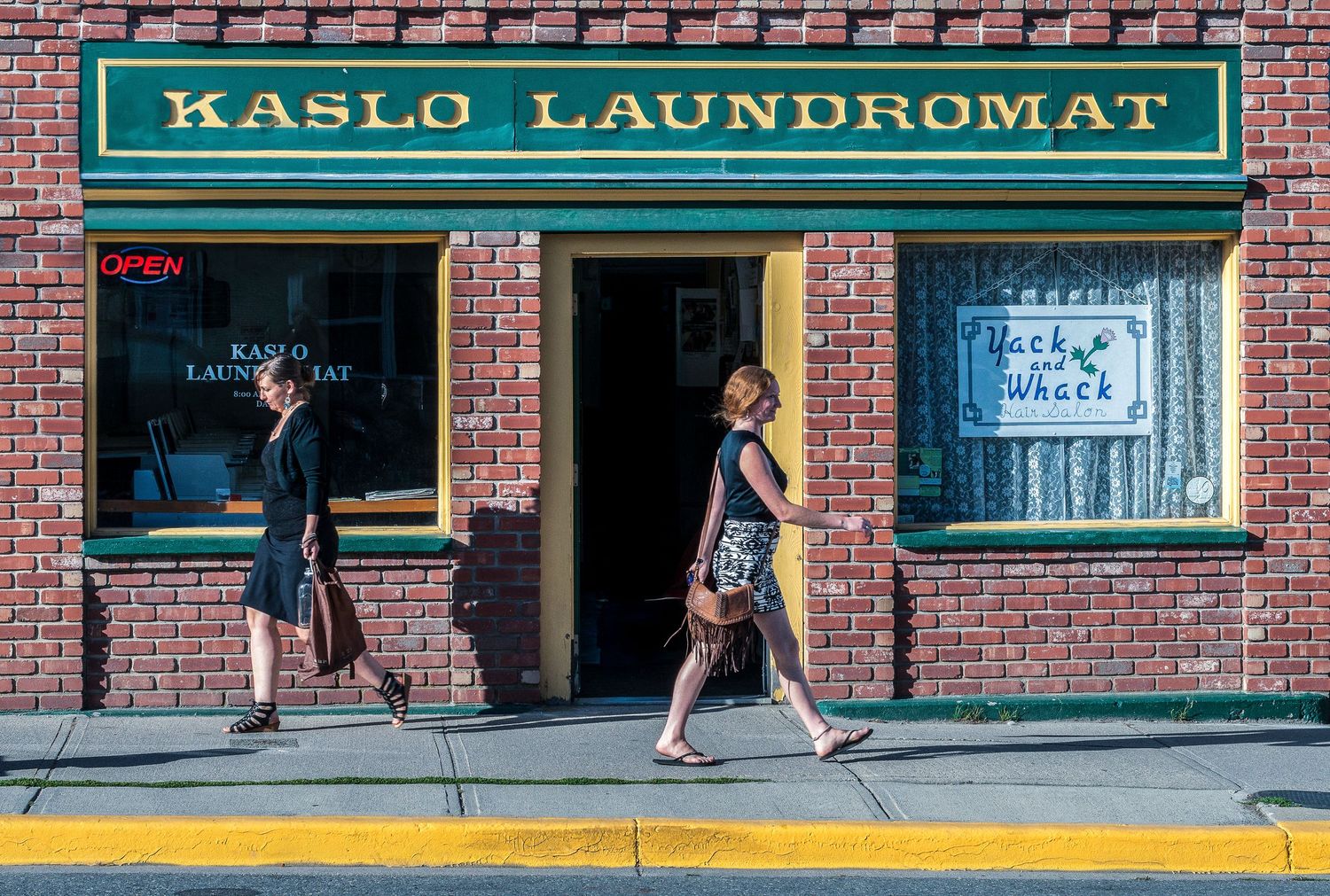 Two people walking past Kaslo Laundromat, a brick storefront with green awning on a sunny day.