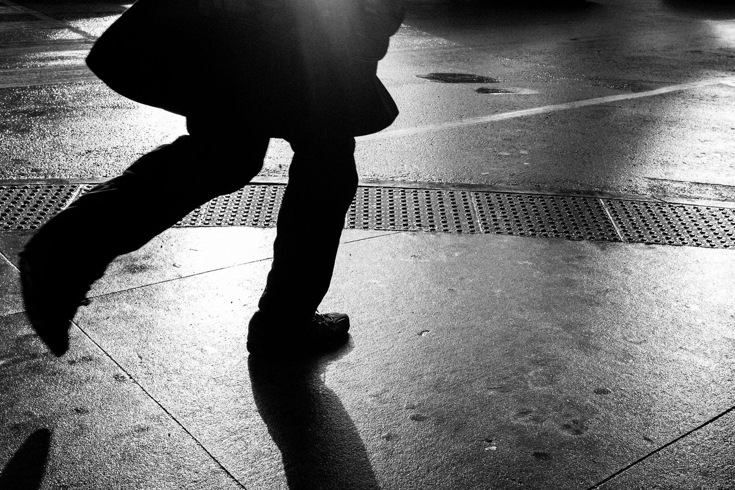 Black and white photo of a person walking on wet pavement, dramatic shadow cast on glistening urban sidewalk.
